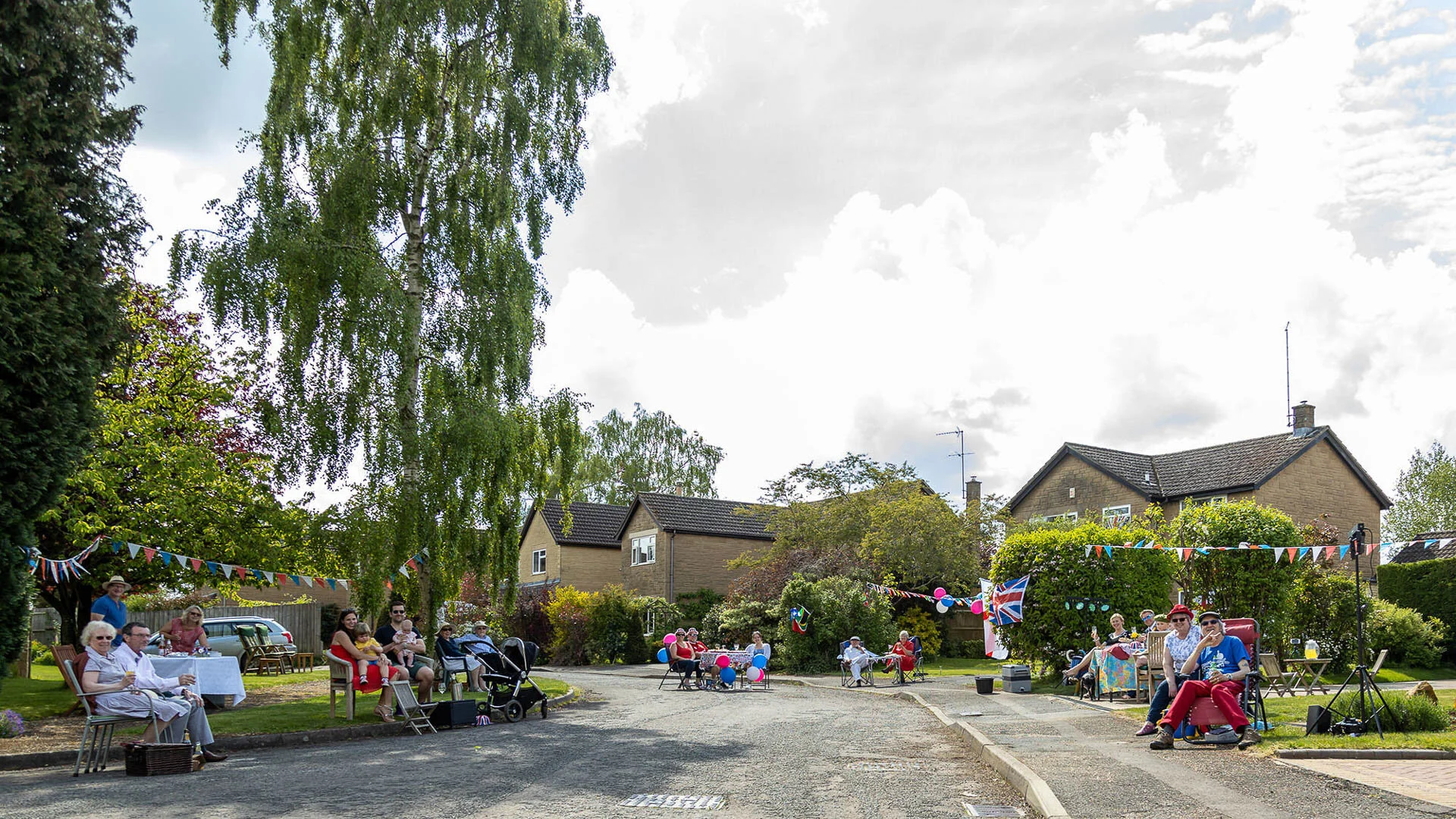 Street party with people sitting on chairs and benches along a residential street. Colorful bunting is hung across the area, and the sky is cloudy. There are parked cars and houses in the background.
