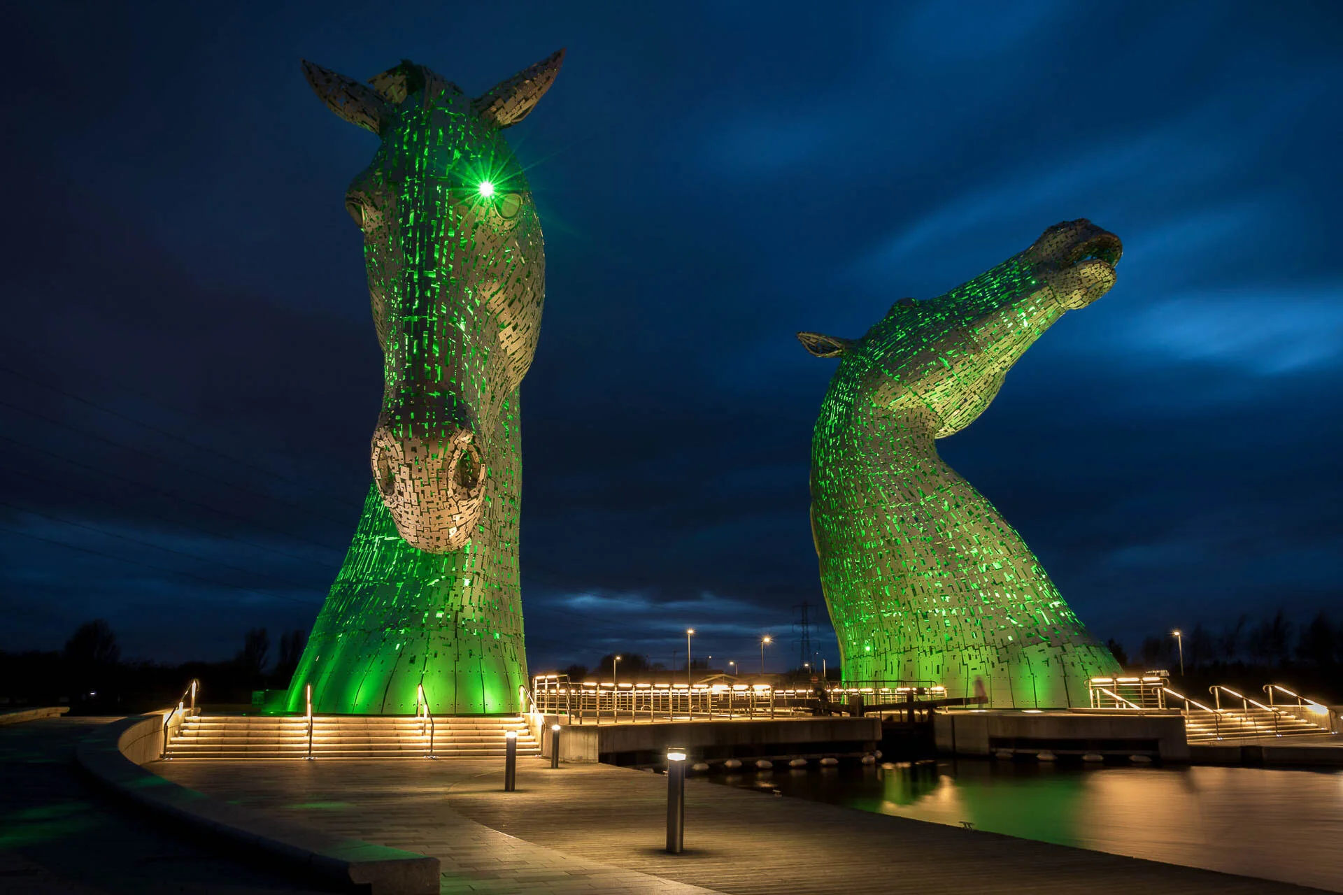Two large horse head sculptures, known as The Kelpies, illuminated with green light against a dark, cloudy sky.