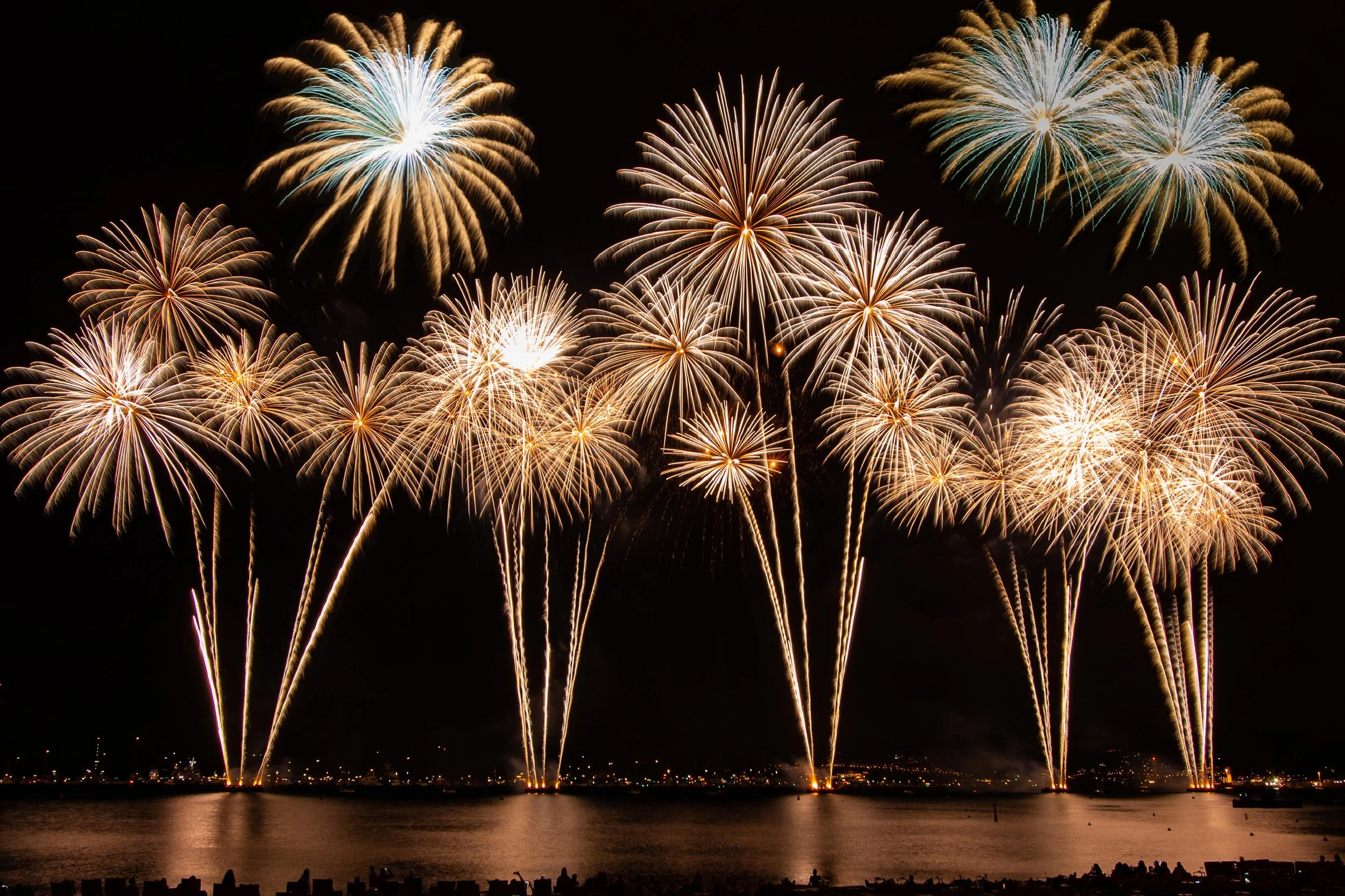 Colorful fireworks exploding over a waterfront at night, reflecting on the water.