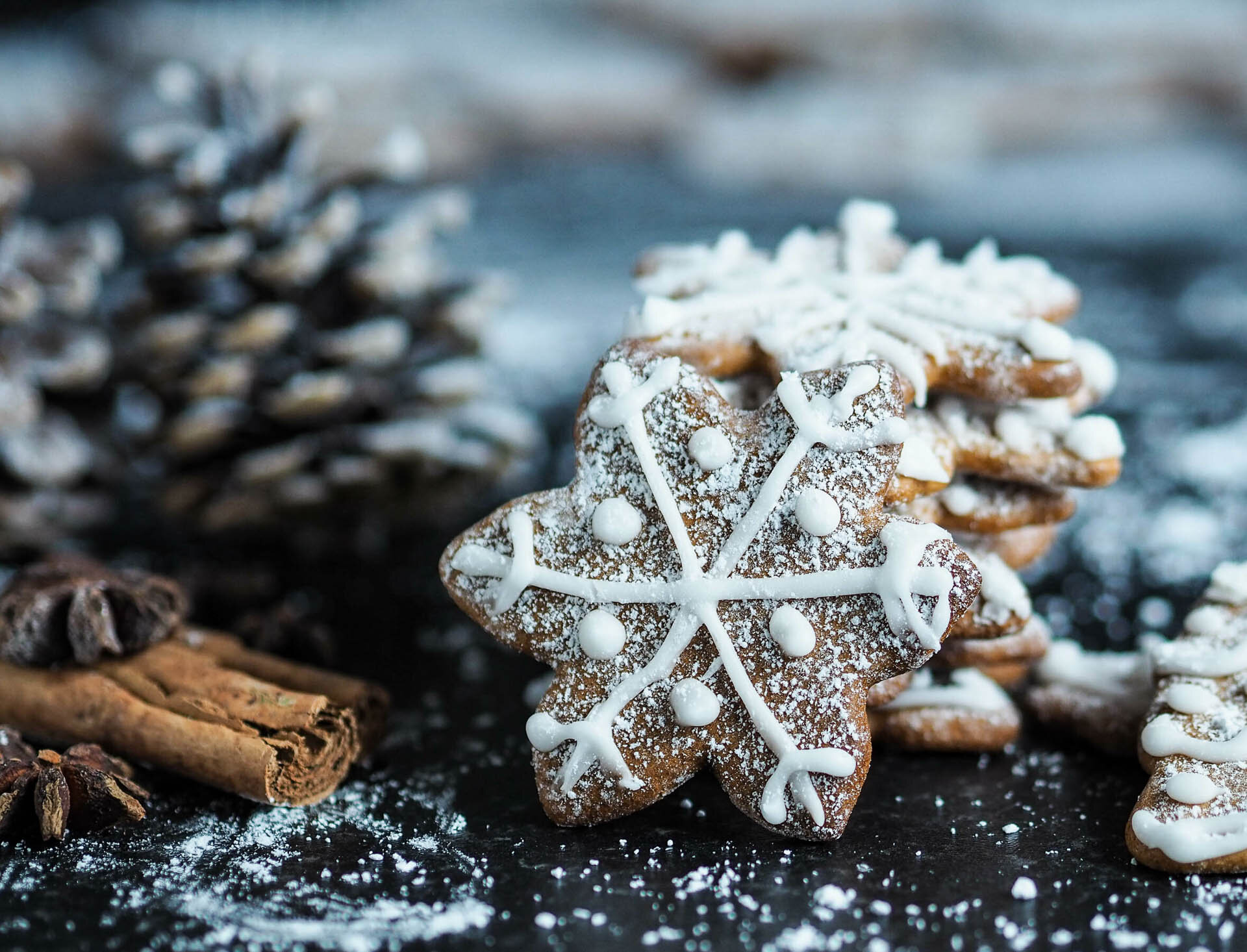 Snowflake-shaped gingerbread cookies with white icing, sprinkled with powdered sugar, surrounded by pinecones and cinnamon sticks on a dark surface.
