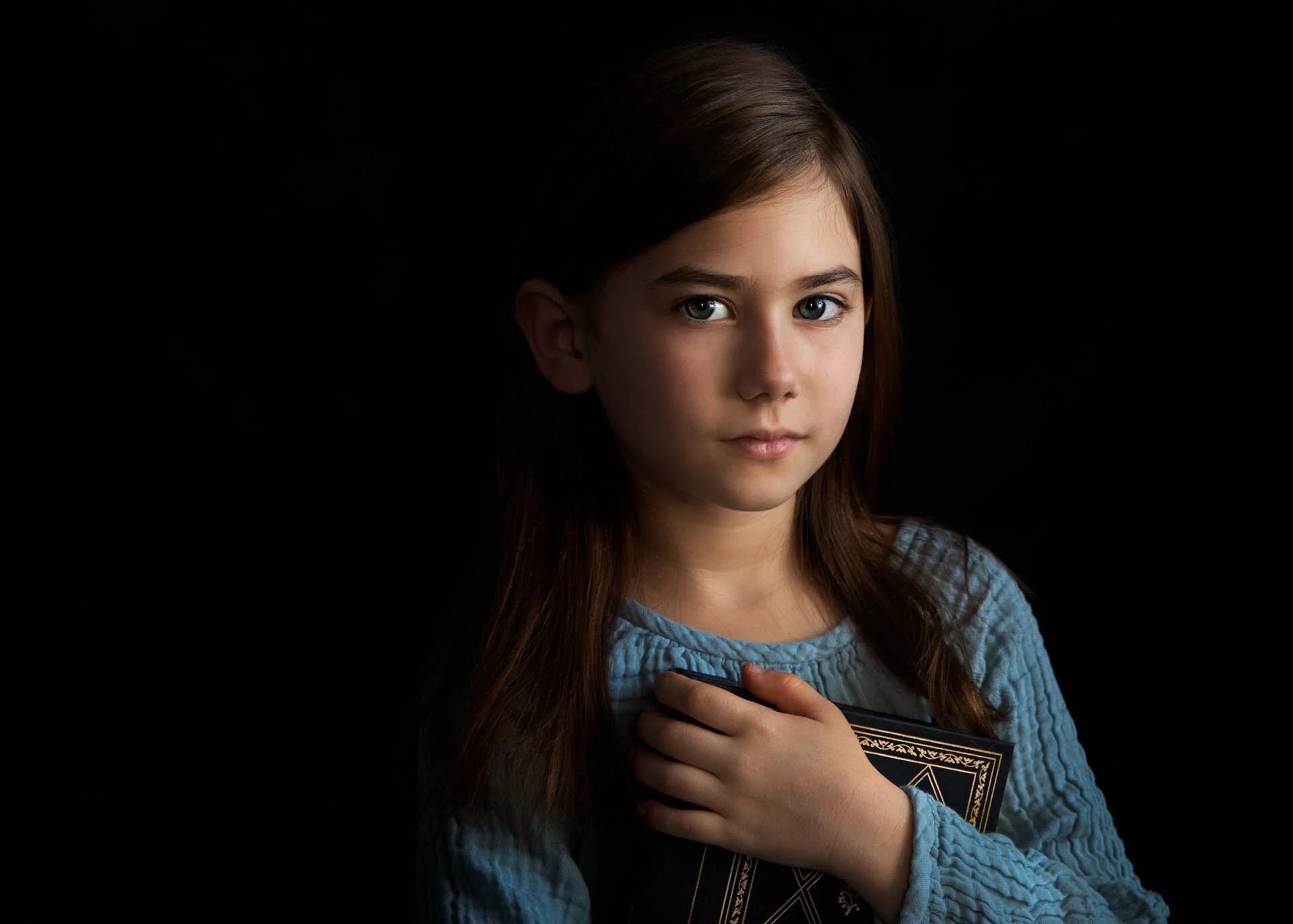 Young girl holding a book with a gold-embossed cover, wearing a blue top, against a dark background.