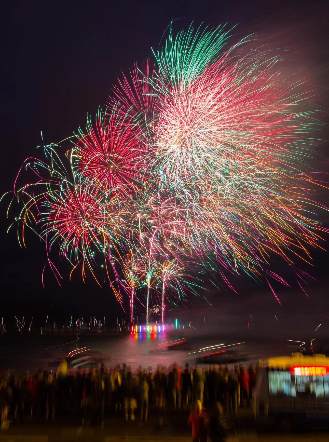 Colorful fireworks display over a body of water at night, with a crowd of people watching in the foreground.