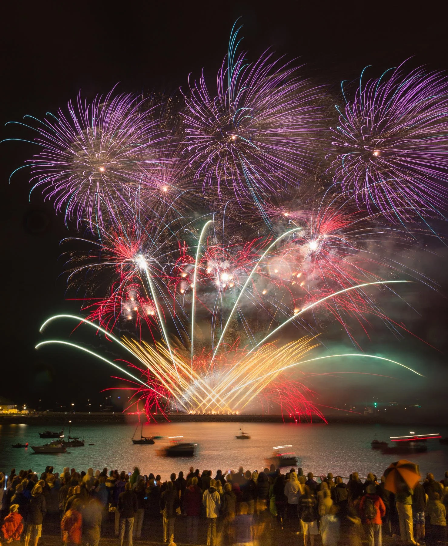 Fireworks display over a body of water with crowds watching from the shore.
