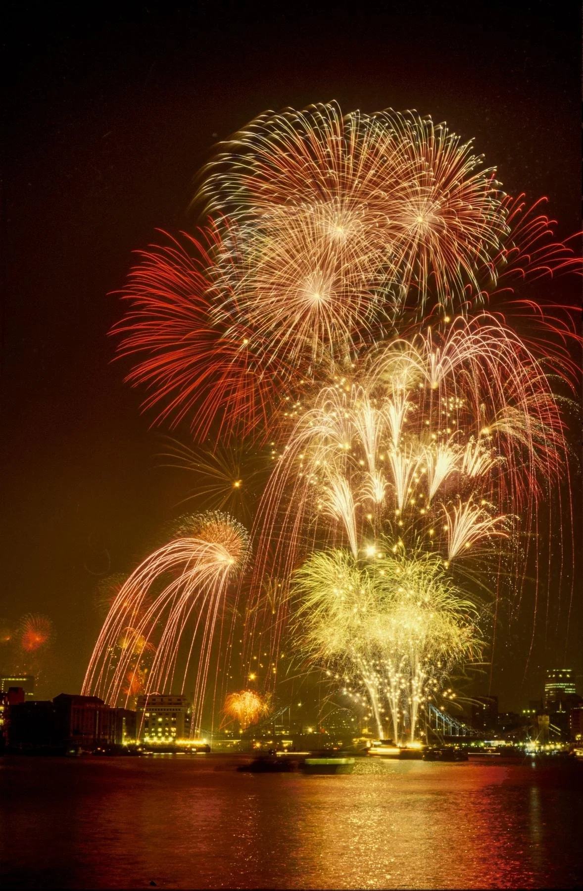 Colorful fireworks display over a city skyline at night with reflections on the water.