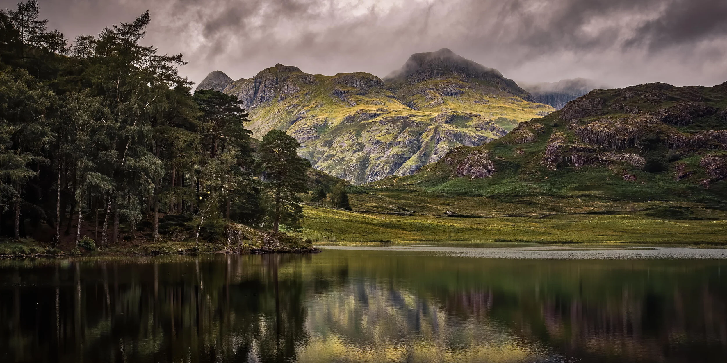 Scenic landscape with a calm lake reflecting lush green hills and rugged mountains under a cloudy sky.
