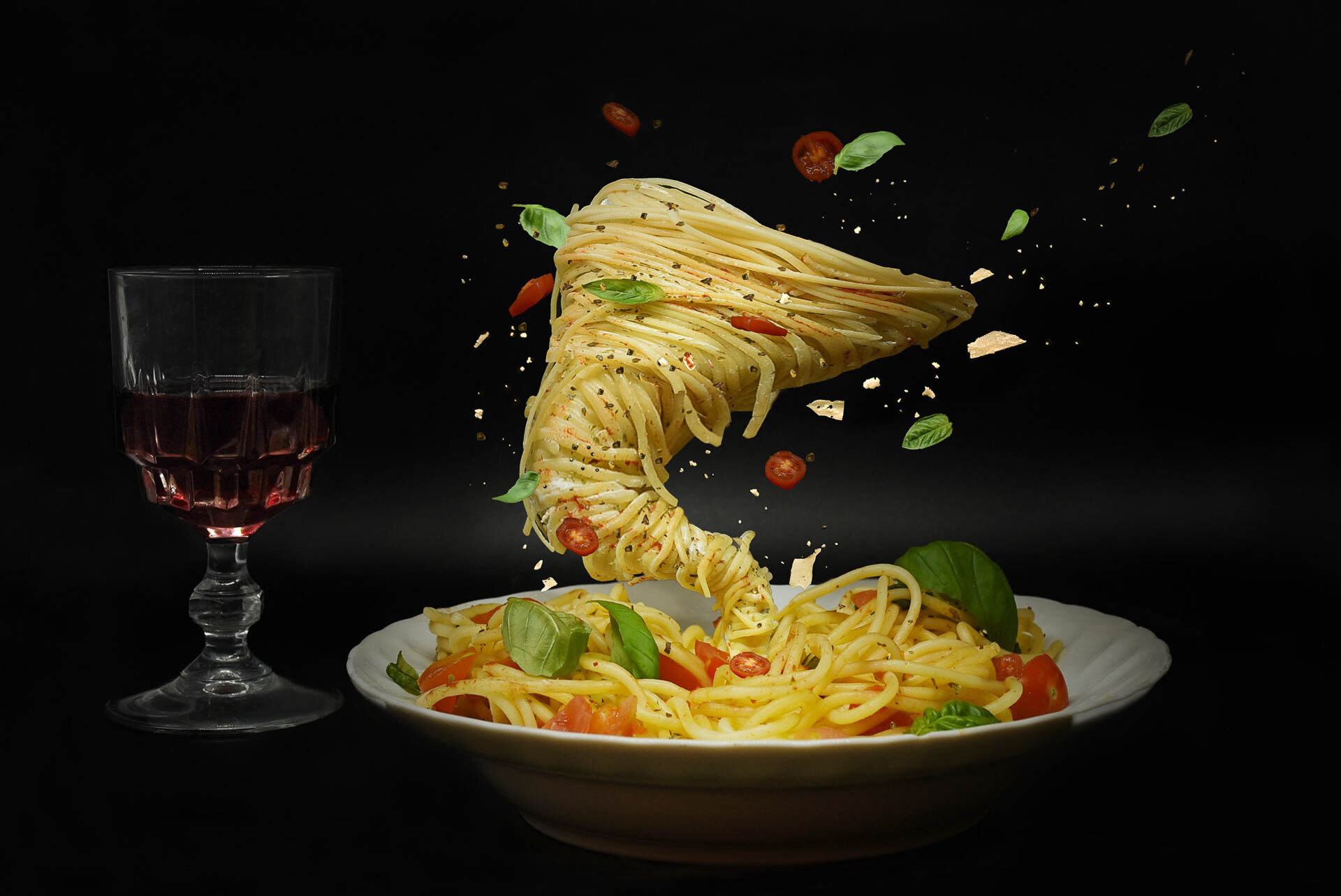 Artistic display of spaghetti and tomatoes swirling upward from a plate with a glass of red wine on the side.