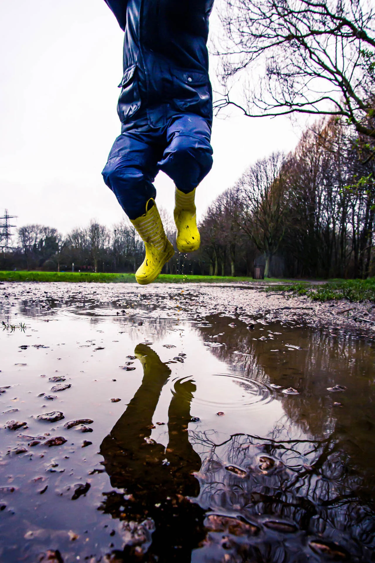 A person jumping in a muddy puddle wearing yellow rain boots and a blue raincoat, with trees in the background.