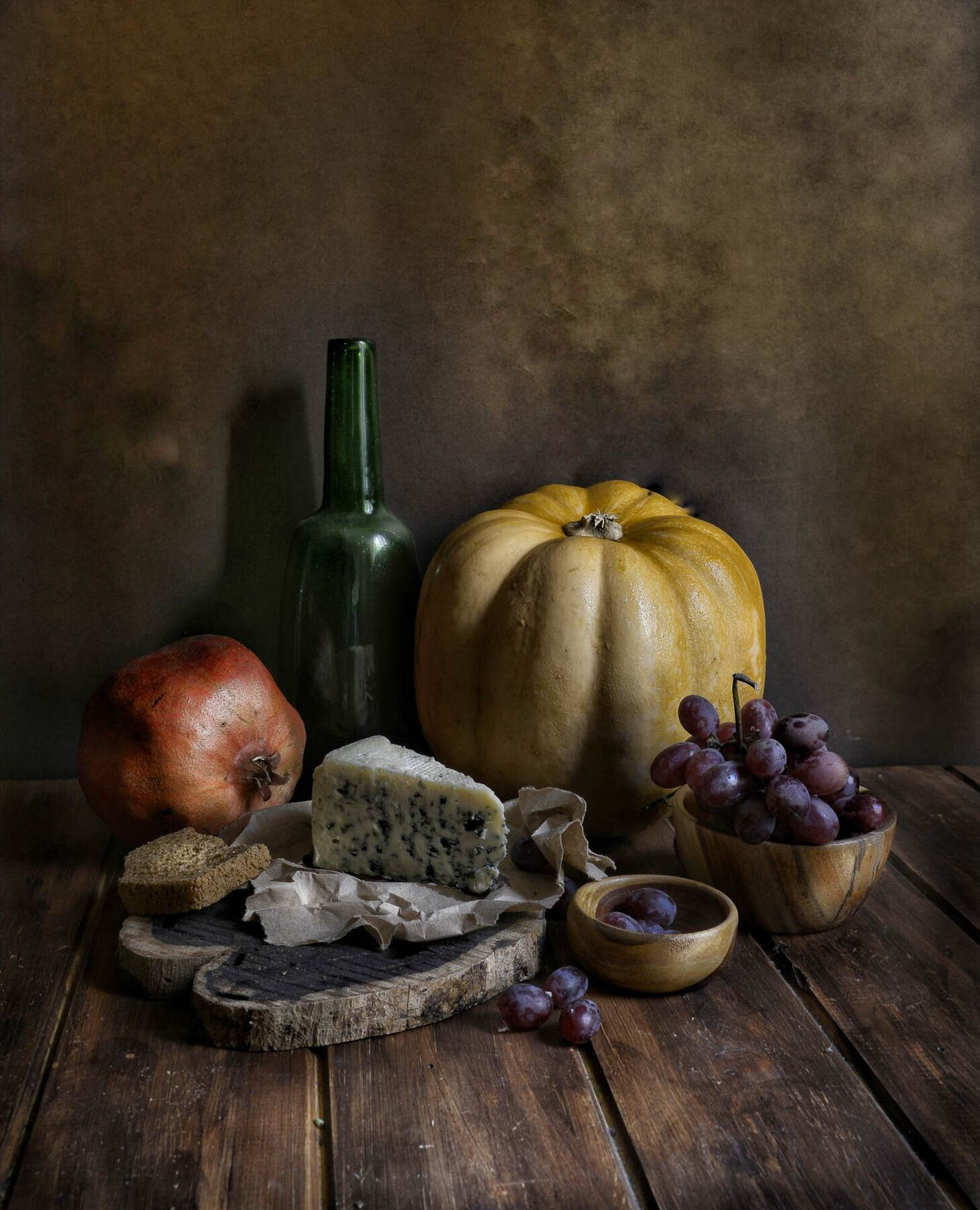 Still life with pumpkin, pomegranate, green bottle, cheese on paper, wooden cutting board, bowl of grapes, and dark bread on wooden table.