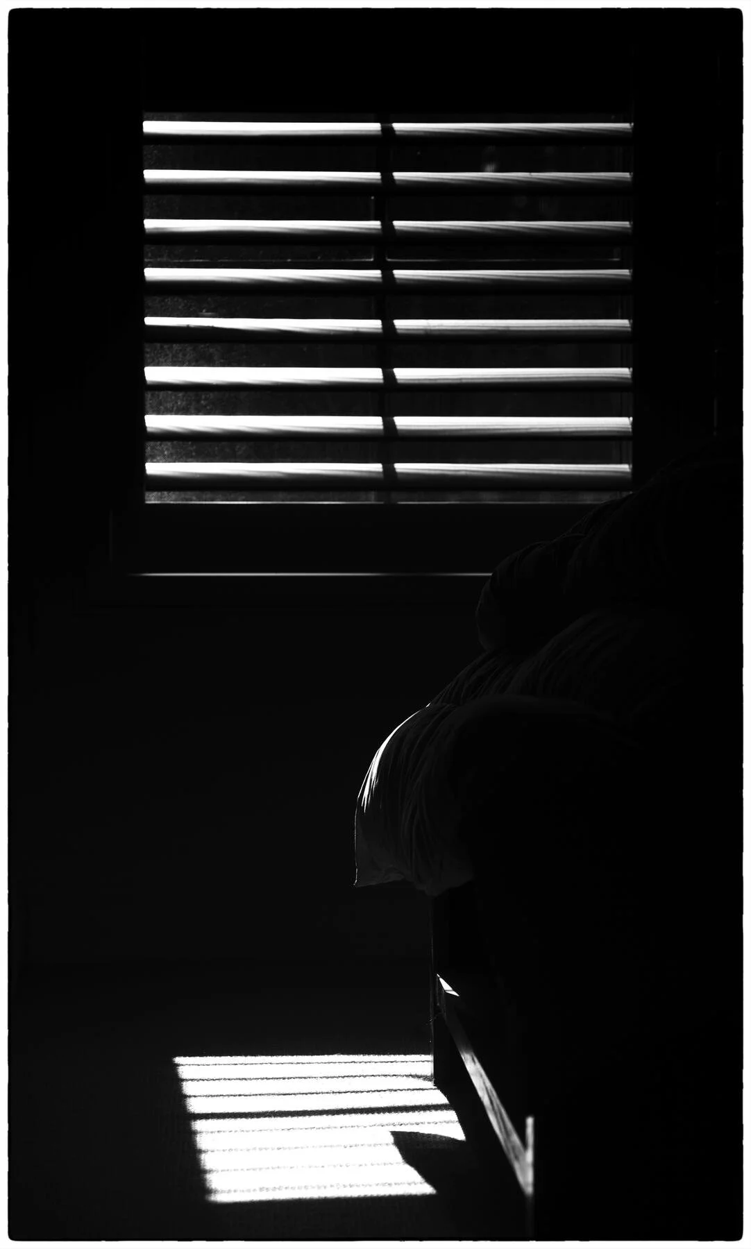 Black and white photo of light streaming through window blinds, casting striped shadows on a bed and floor.