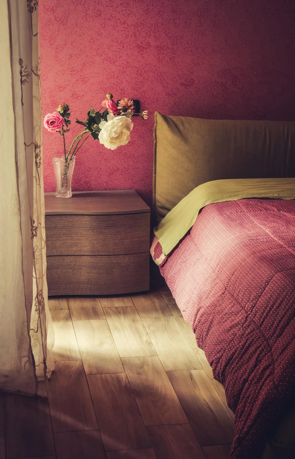 Cozy bedroom with pink walls, wooden nightstand, flowers in a vase, and a bed with a red comforter.