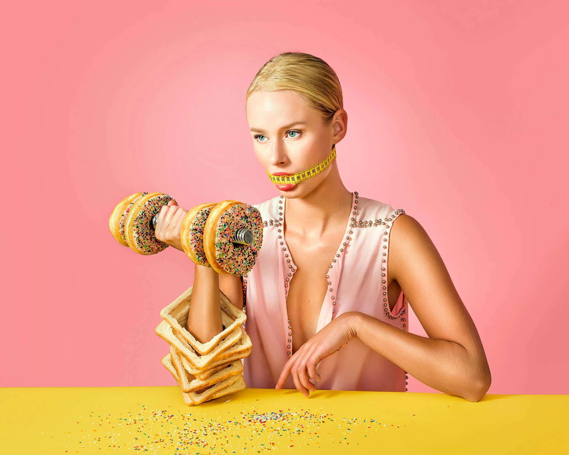 A woman with a measuring tape wrapped around her mouth lifts a dumbbell made to look like two sprinkled donuts. Her arm is stacked with slices of bread. The background is pink, and she is leaning on a yellow table sprinkled with colorful bits.