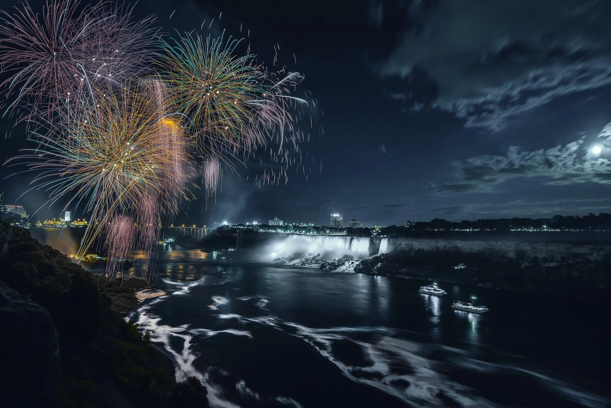 Fireworks over Niagara Falls at night with illuminated waterfalls and boats on the river.