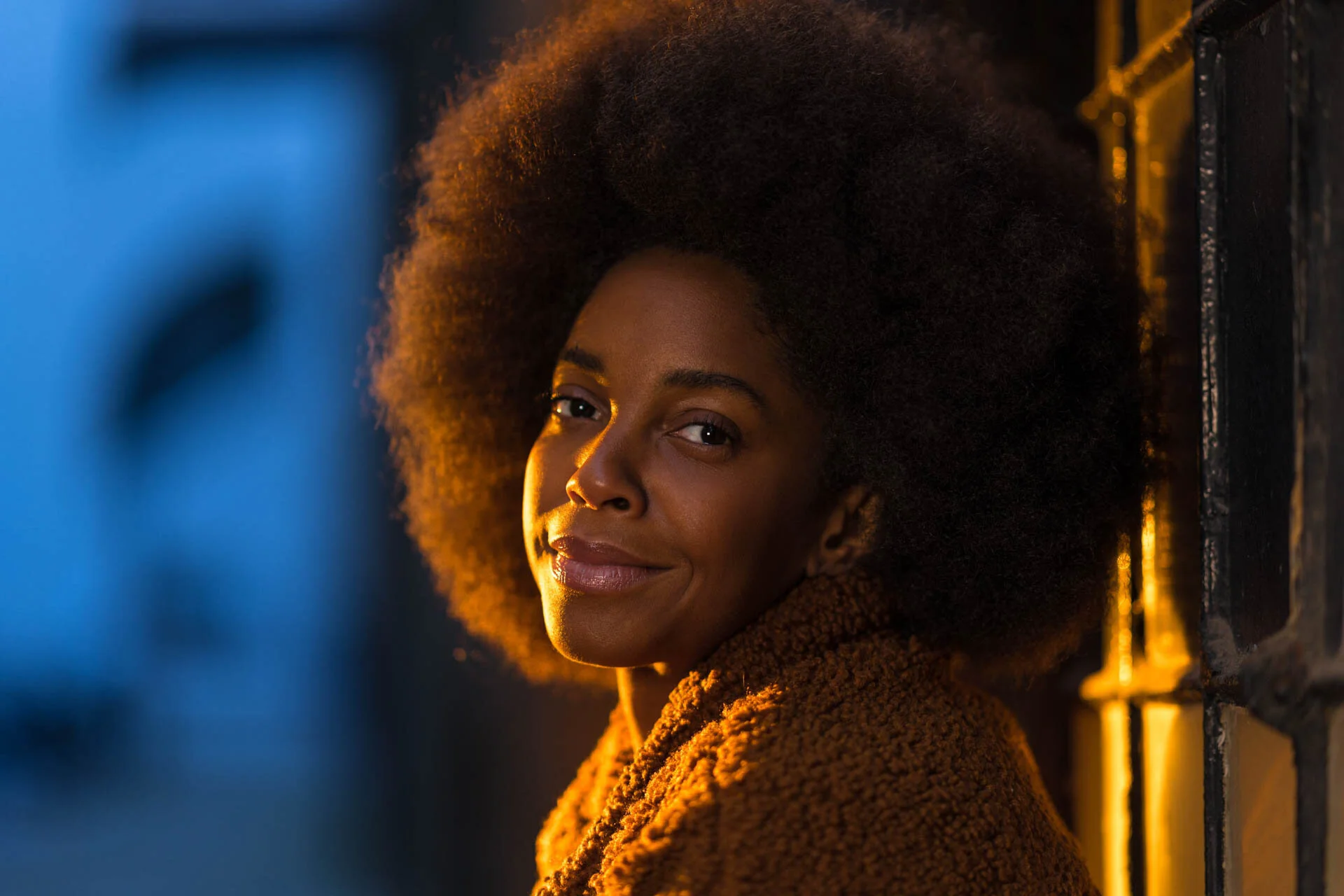 Portrait of a woman with an afro hairstyle and brown coat, leaning against a wall, with warm lighting.
