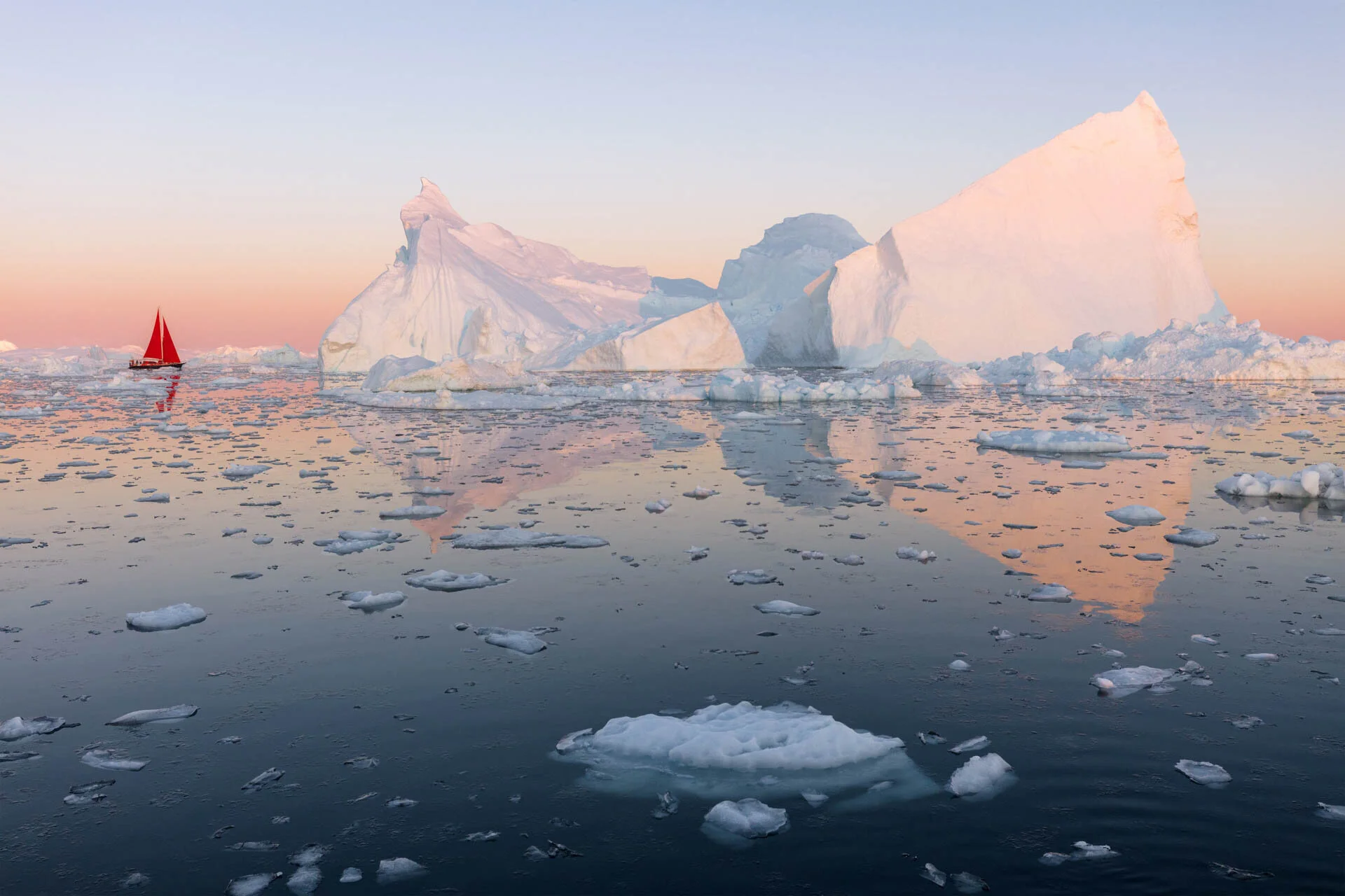 Red-sailed boat near large icebergs in a calm, icy sea under a pink and orange sunset.