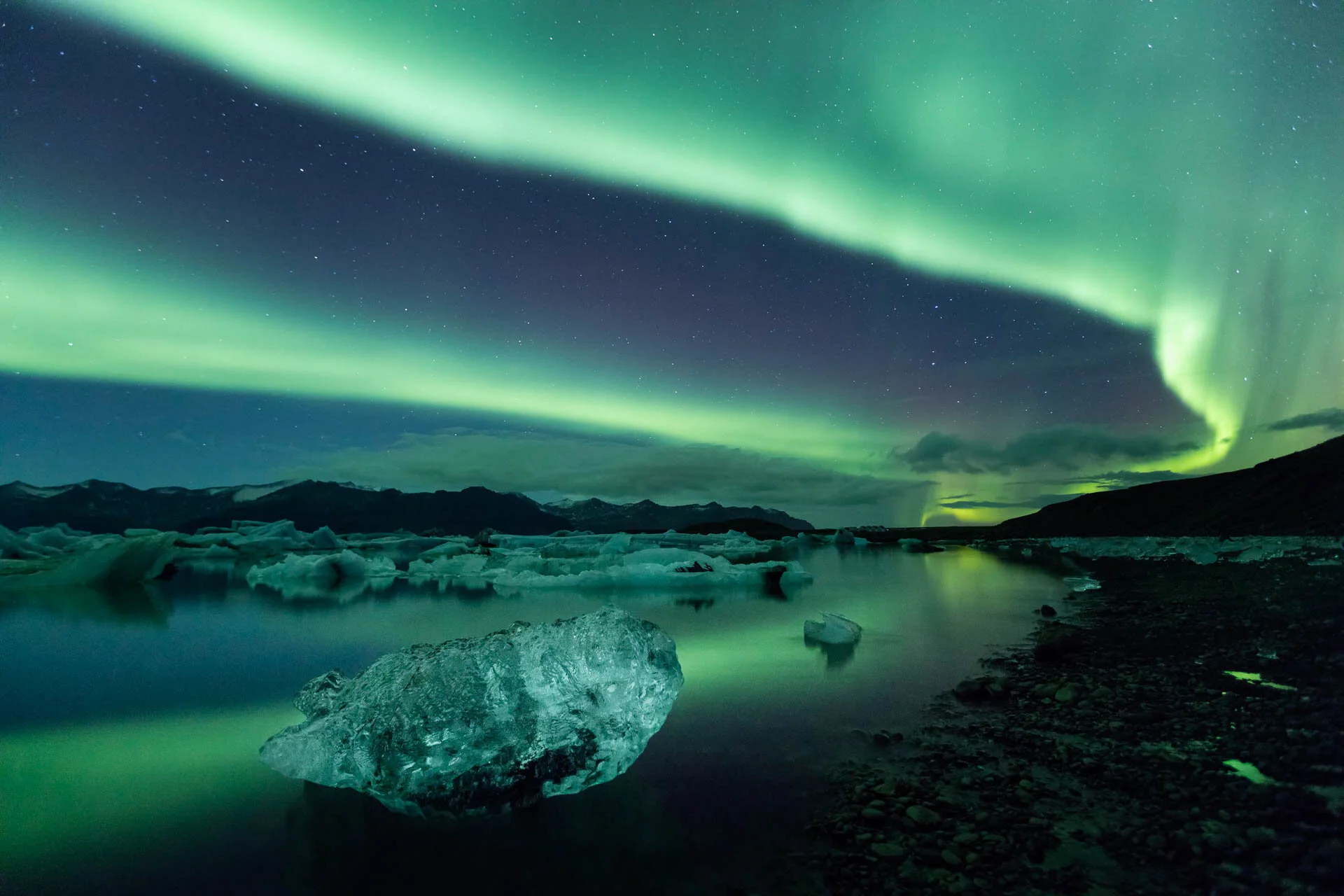 Northern lights over a glacial lagoon with icebergs and mountains.