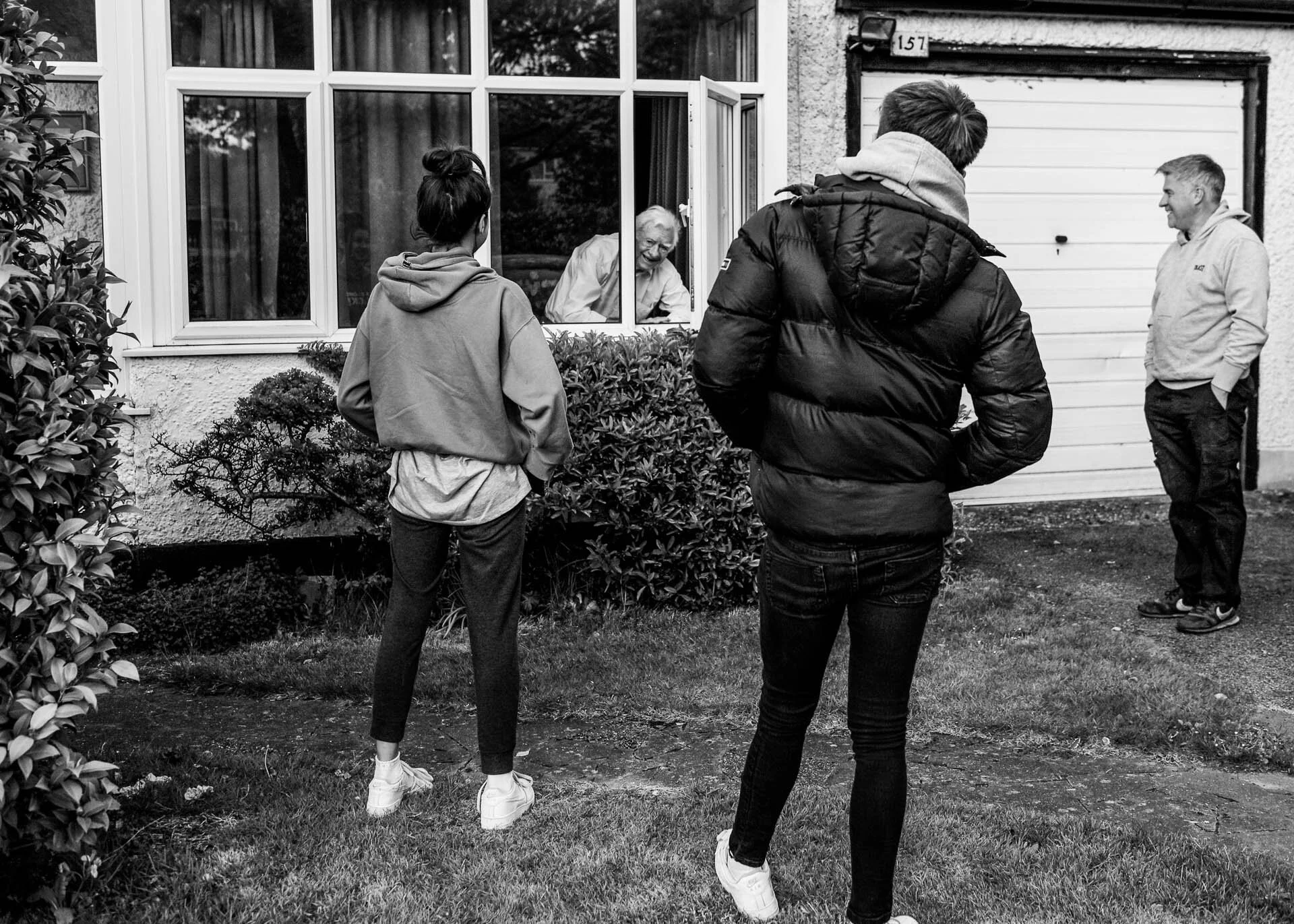 Black and white photo of two people standing outside near a window talking with an elderly person indoors, with another man standing nearby, and a garage door in the background.