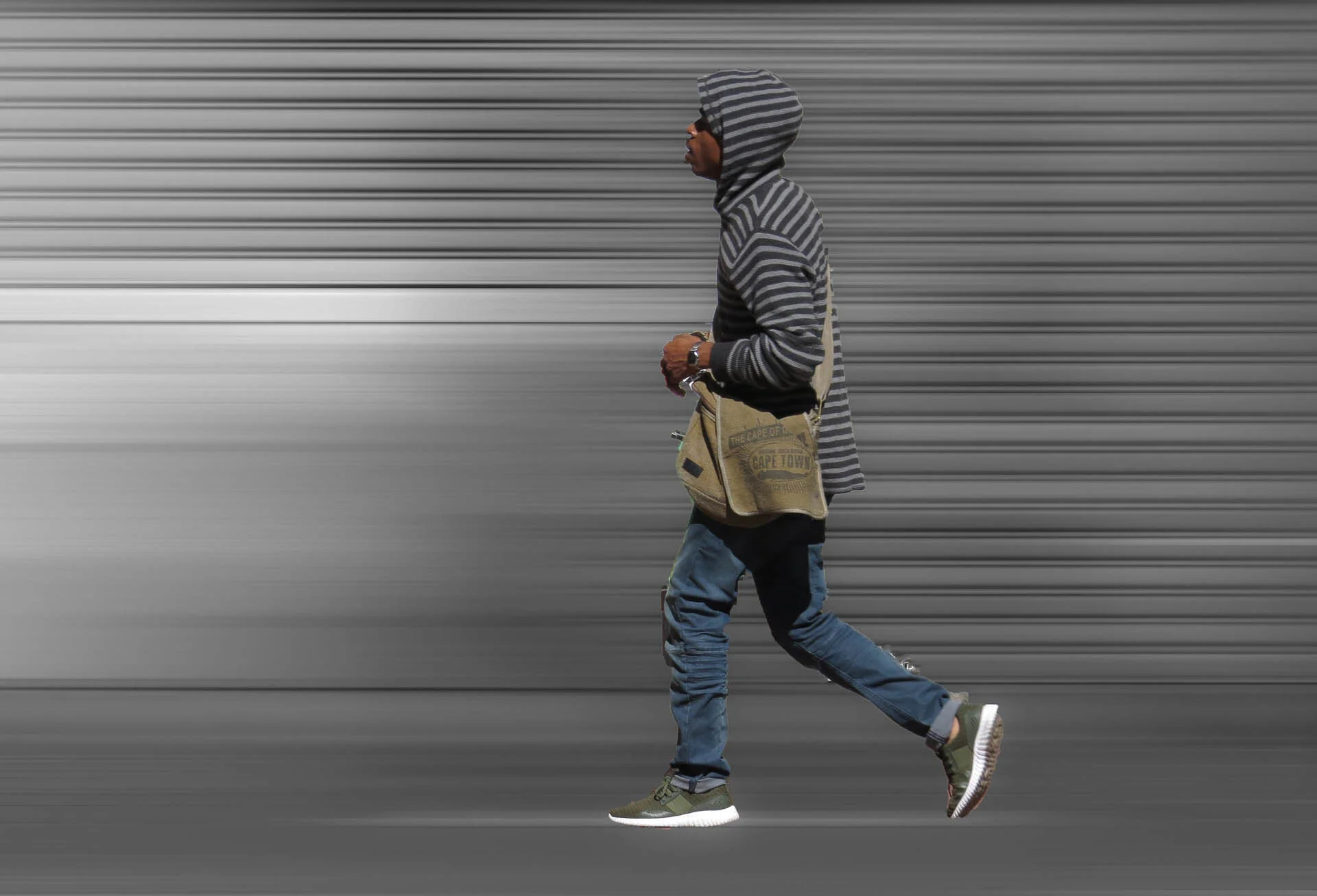 Person wearing a striped hoodie walking with a messenger bag against a blurred industrial background.