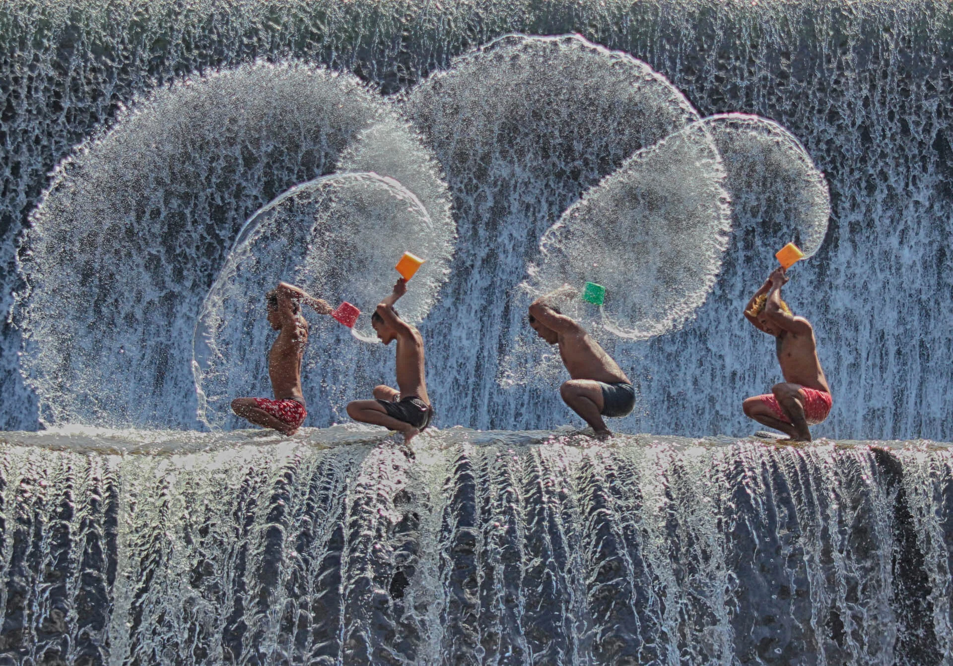 Four people crouching near a waterfall, splashing water with colorful buckets, creating arcs in the air.