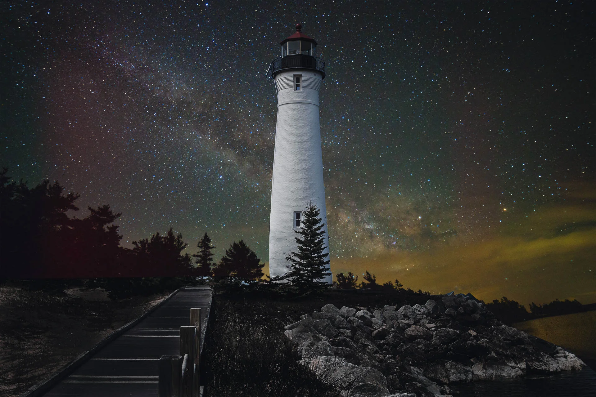 Lighthouse under a starry night sky with Milky Way, surrounded by trees and rocks, ocean in background.