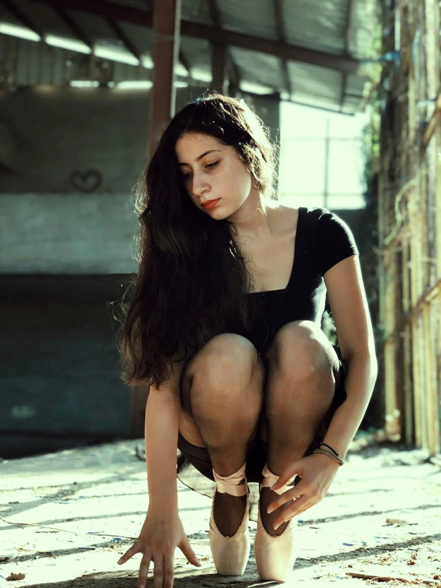 A woman in a black dress crouching gracefully on a sunlit floor, wearing ballet pointe shoes, with a thoughtful expression in an industrial setting.