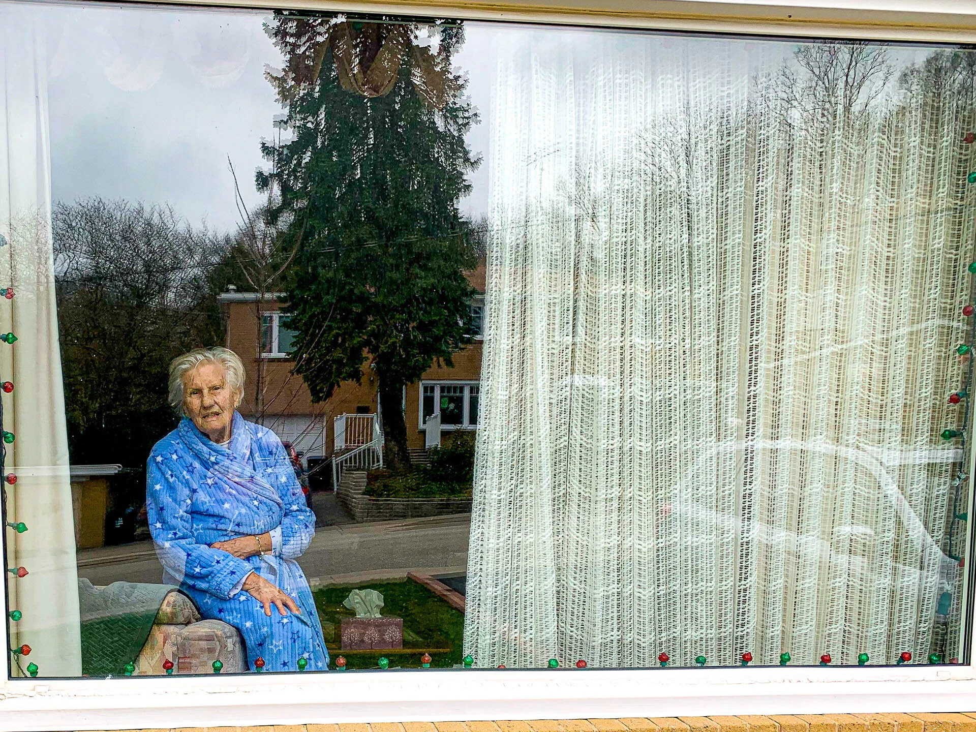An elderly person in a blue robe sitting inside a house, seen through a window with white curtains and reflections of trees and a car outside.