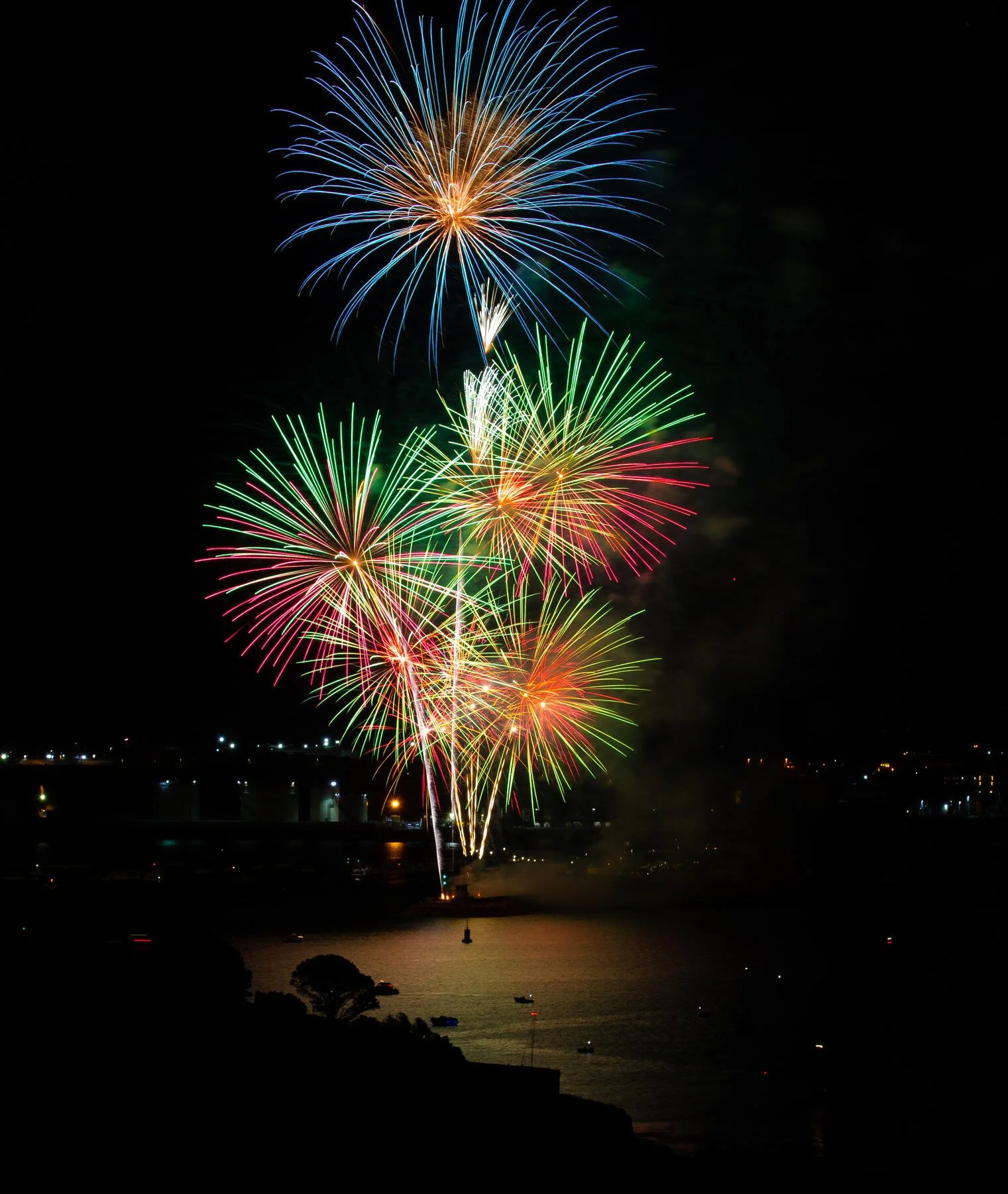 Colorful fireworks display over a body of water at night, with boats and city lights in the background.