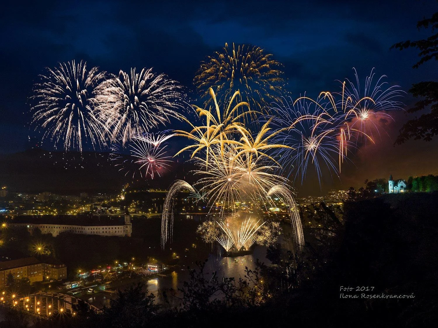 Fireworks display over a city at night with a river and buildings illuminated by lights.