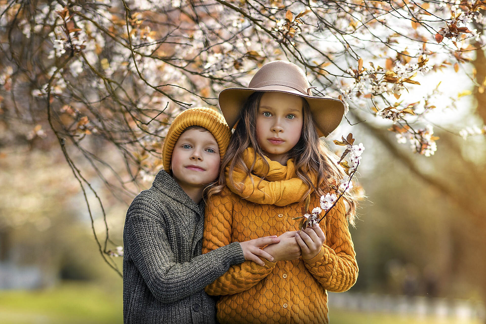 Two children in autumn clothing, a boy wearing a yellow beanie and a gray sweater, and a girl with long hair in a wide-brimmed hat and yellow scarf, standing under a blossoming tree.