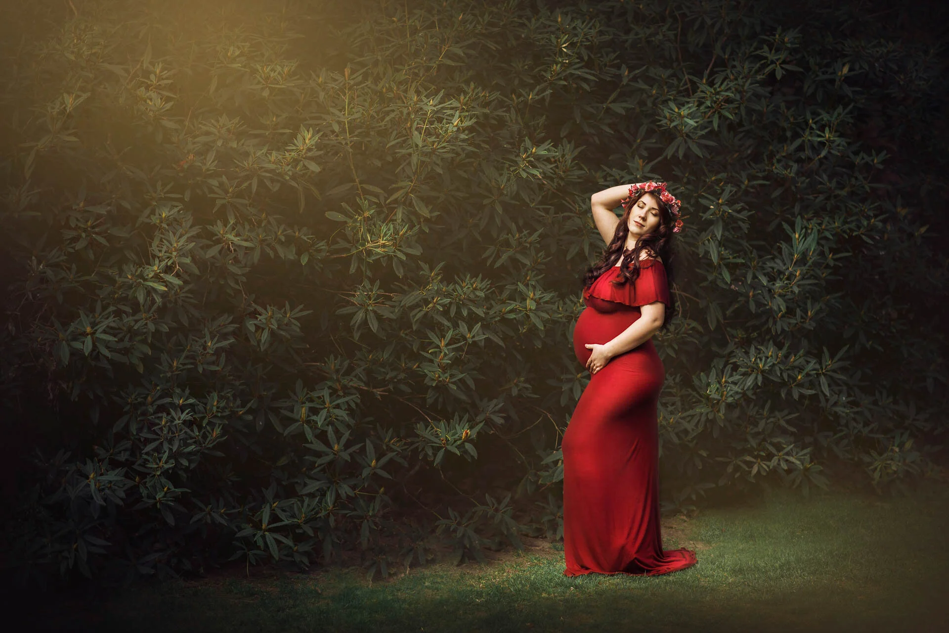 Pregnant woman in a red dress posing outdoors against a backdrop of lush green foliage, wearing a flower crown, with soft lighting creating a serene atmosphere.
