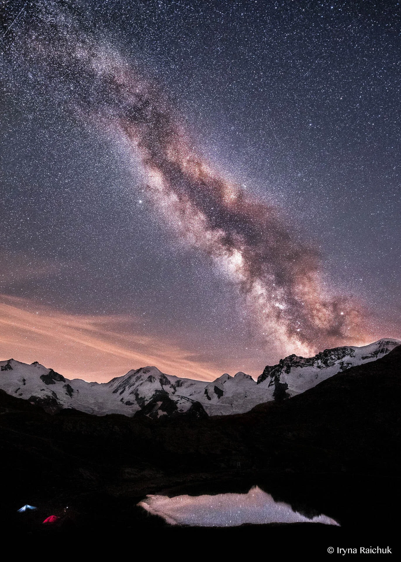Milky Way over snow-covered mountains and a reflective lake at night with stars visible in the sky.
