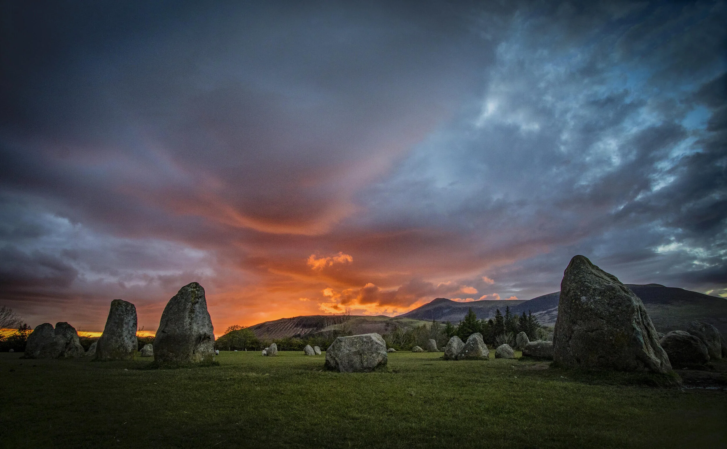 Stone circle on a grassy field with a dramatic sunset sky, featuring orange and blue clouds, and mountains in the background.