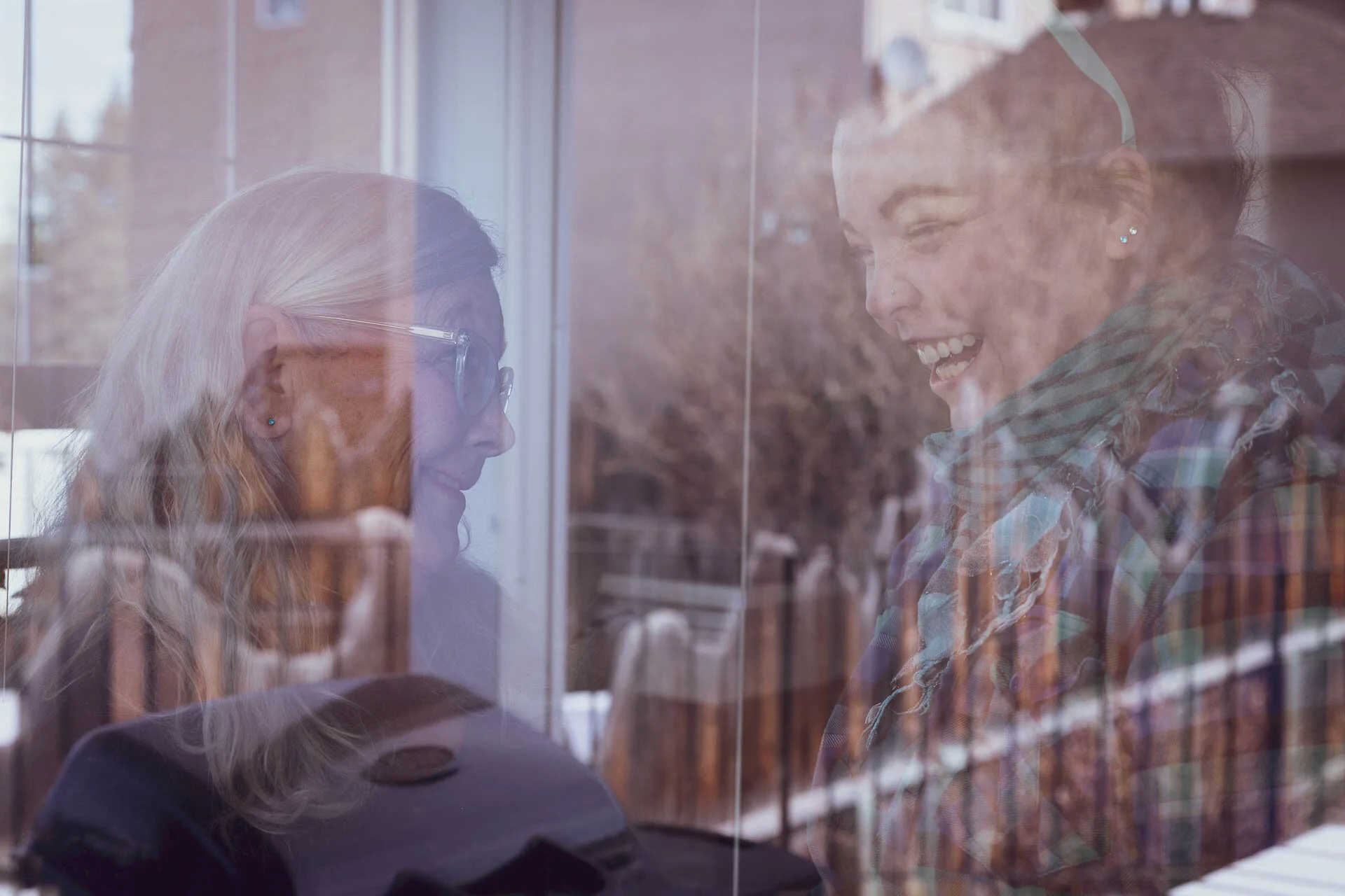 Two people smiling and talking, observed through a reflective window with blurred outdoor scenery in the background.