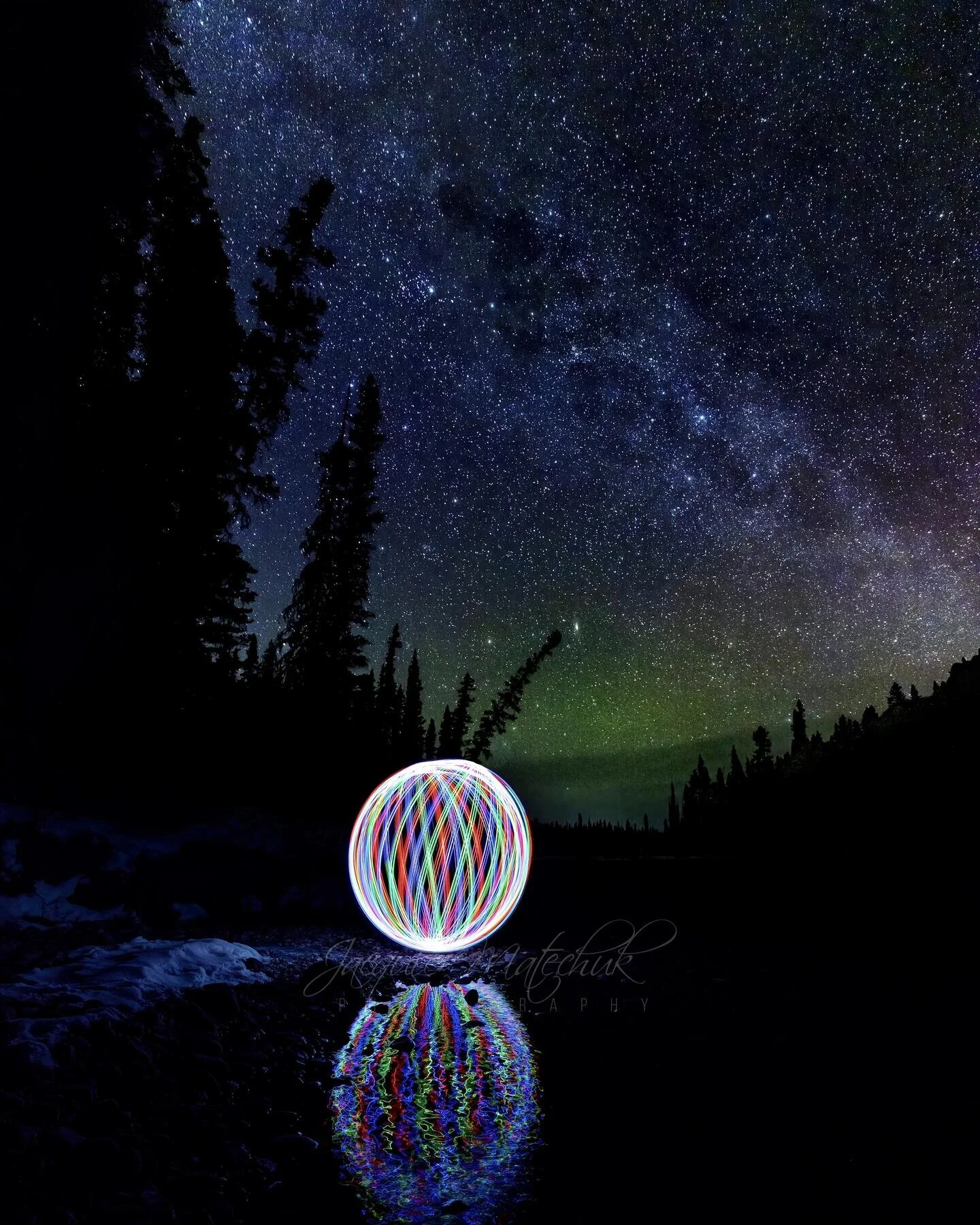 Night sky with stars and Milky Way, silhouetted trees, and a colorful light orb reflected in water.