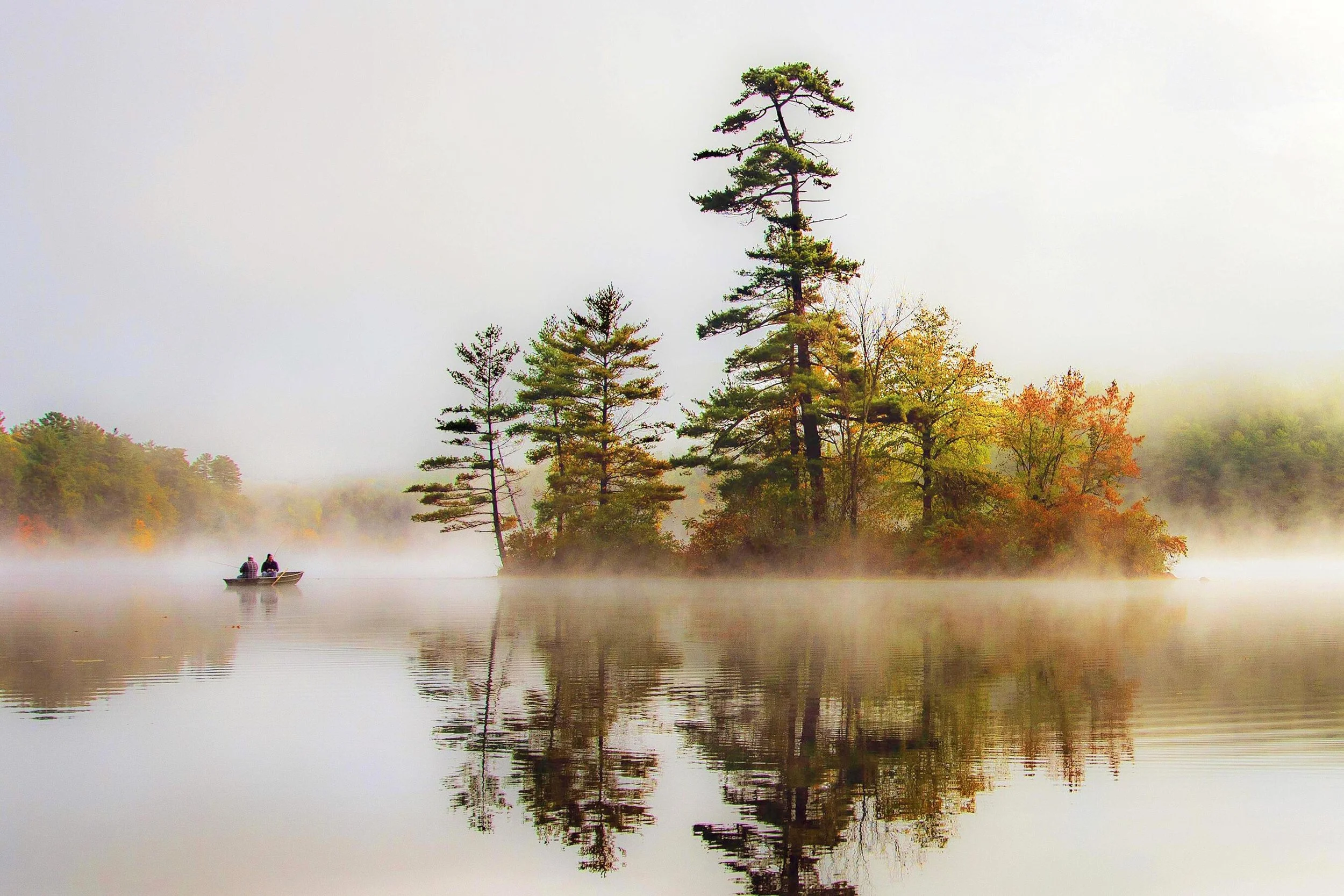 Small boat with two people on misty lake reflecting trees in autumn.