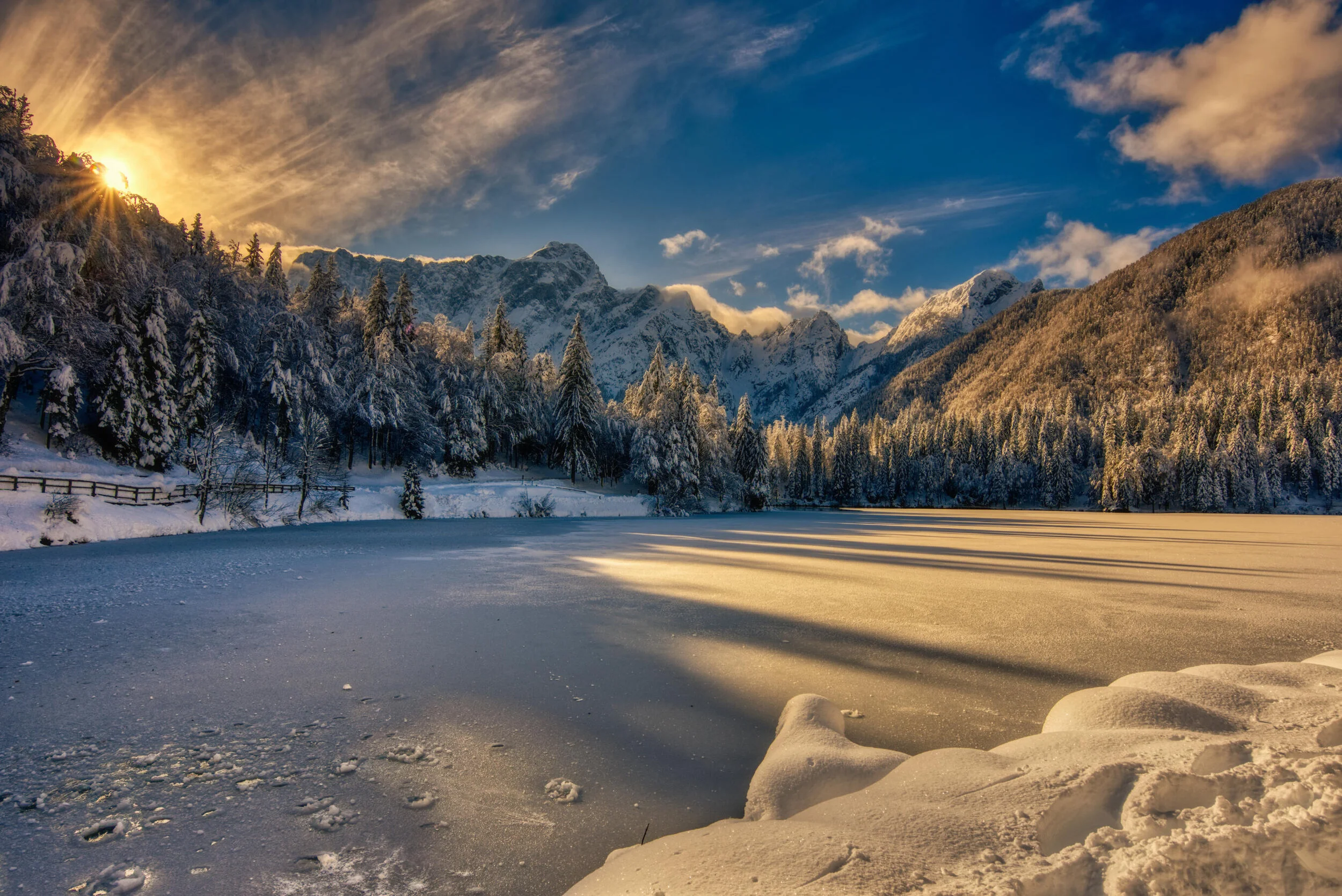 Snow-covered landscape with a frozen lake, surrounded by trees and mountains at sunset.