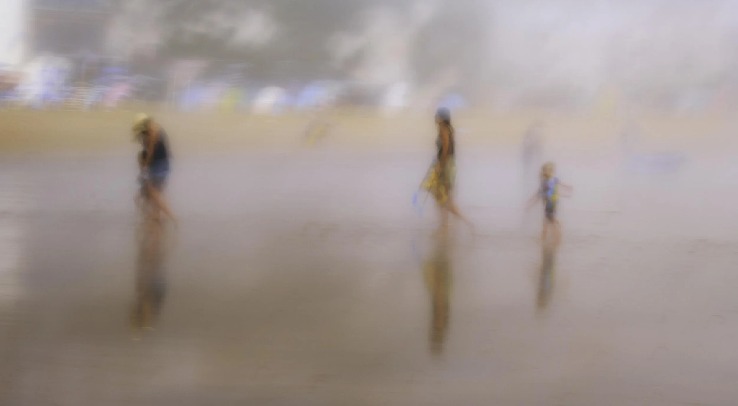 Blurred beach scene with people walking on wet sand, reflected in the water, and misty background.