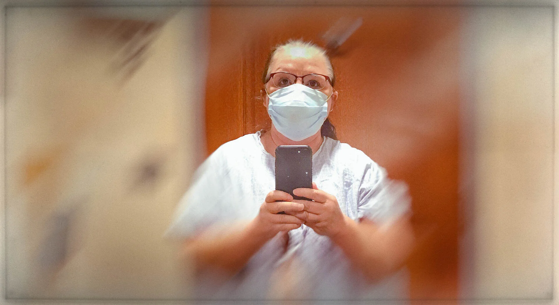 Person wearing a face mask and glasses taking a selfie with a smartphone in a mirror, background is blurred.