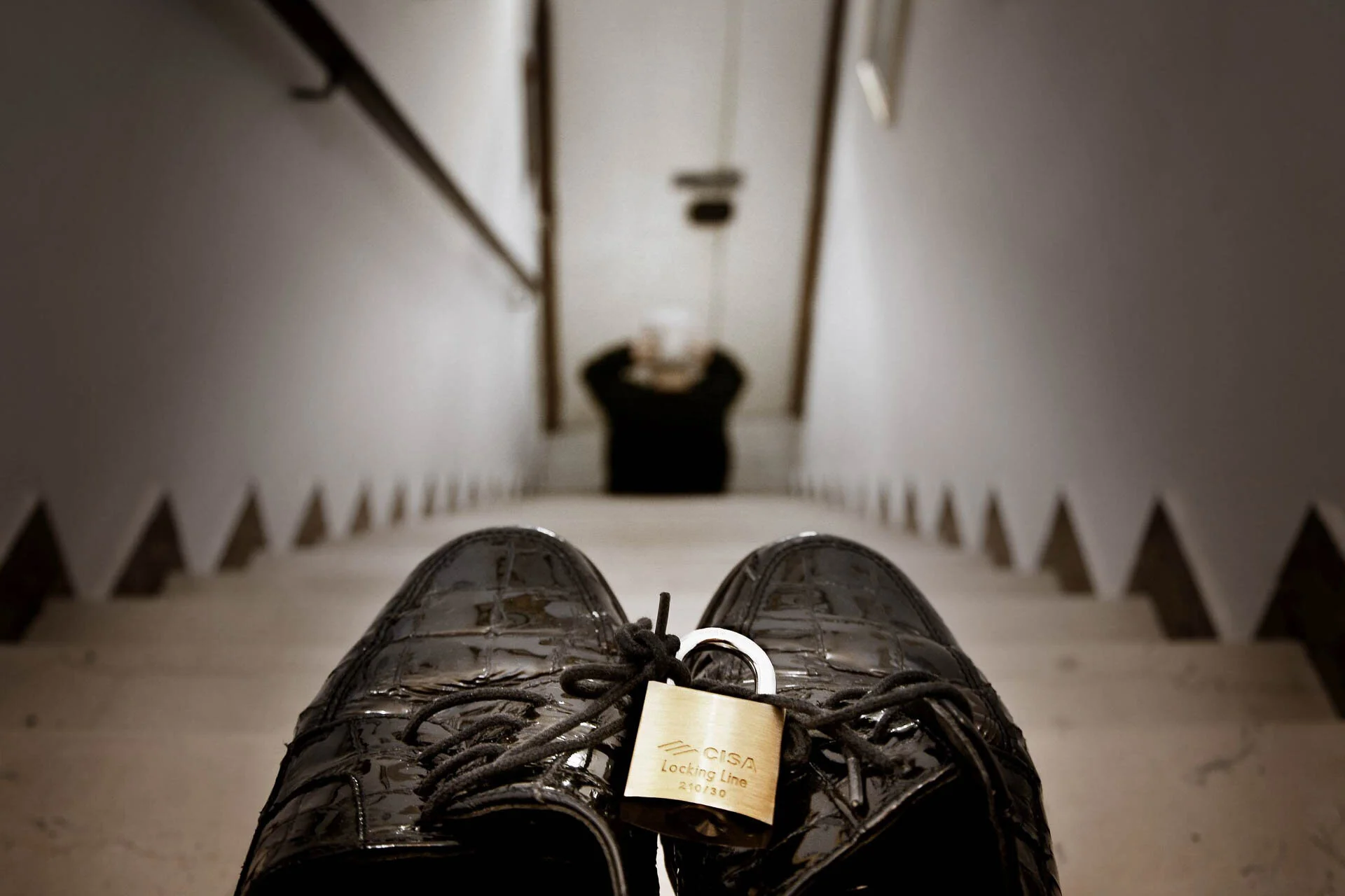 Pair of black shoes with a padlock on the laces placed on stairs, with a person sitting at the bottom.