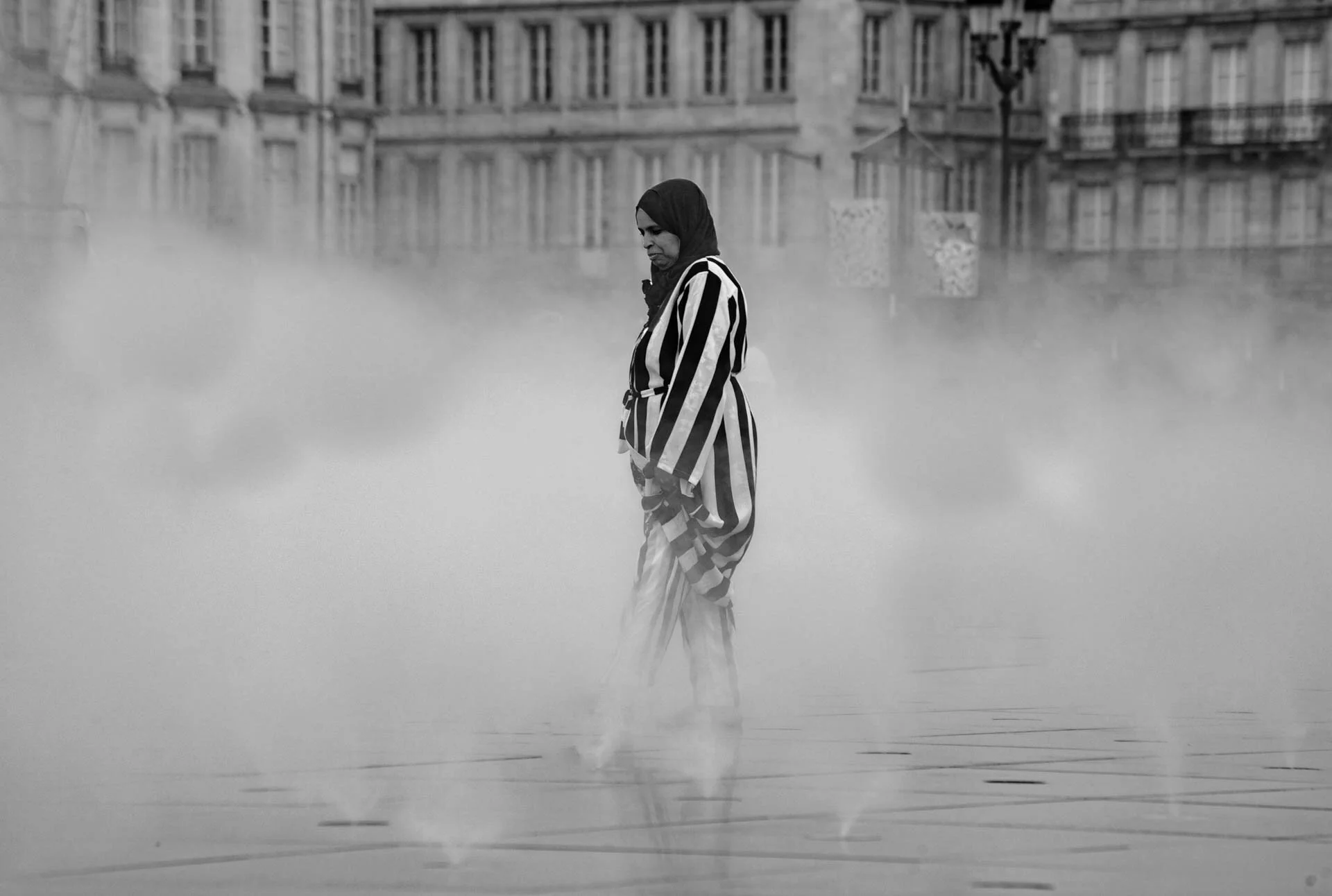 A person in a striped outfit standing amidst fog in an urban setting, with blurred buildings in the background.