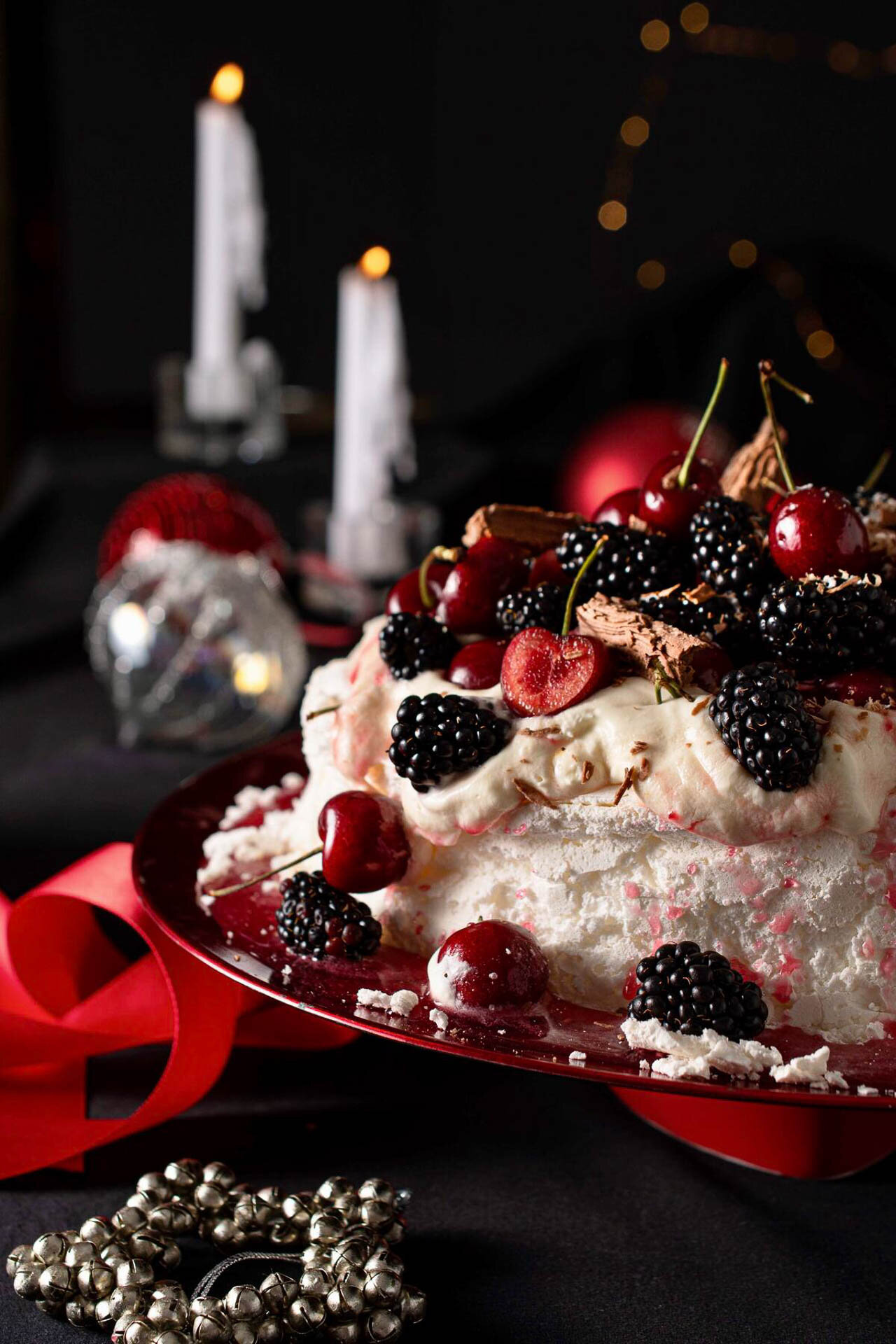 Festive dessert with whipped cream, cherries, blackberries, and chocolate shavings on a red plate, surrounded by silver bells and lit candles.