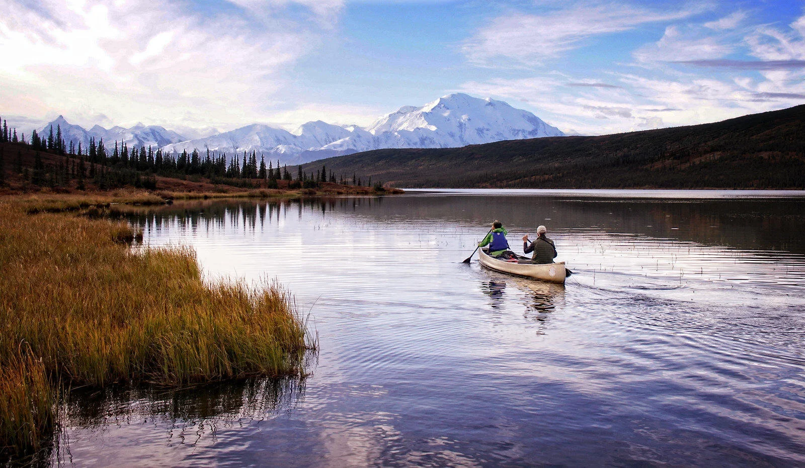 Two people canoeing on a lake surrounded by mountains and pine trees under a cloudy sky.