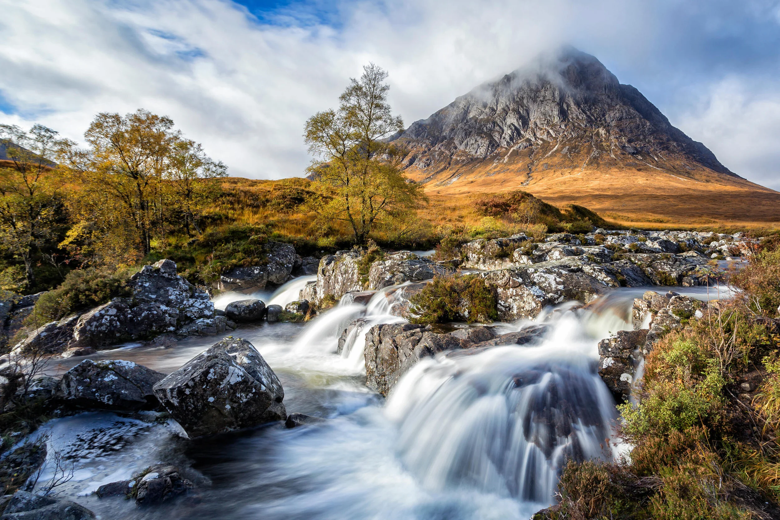 Scenic landscape of a mountain partially shrouded in clouds, with a cascading stream over rocky terrain and autumn-colored trees in the foreground, under a cloudy blue sky.