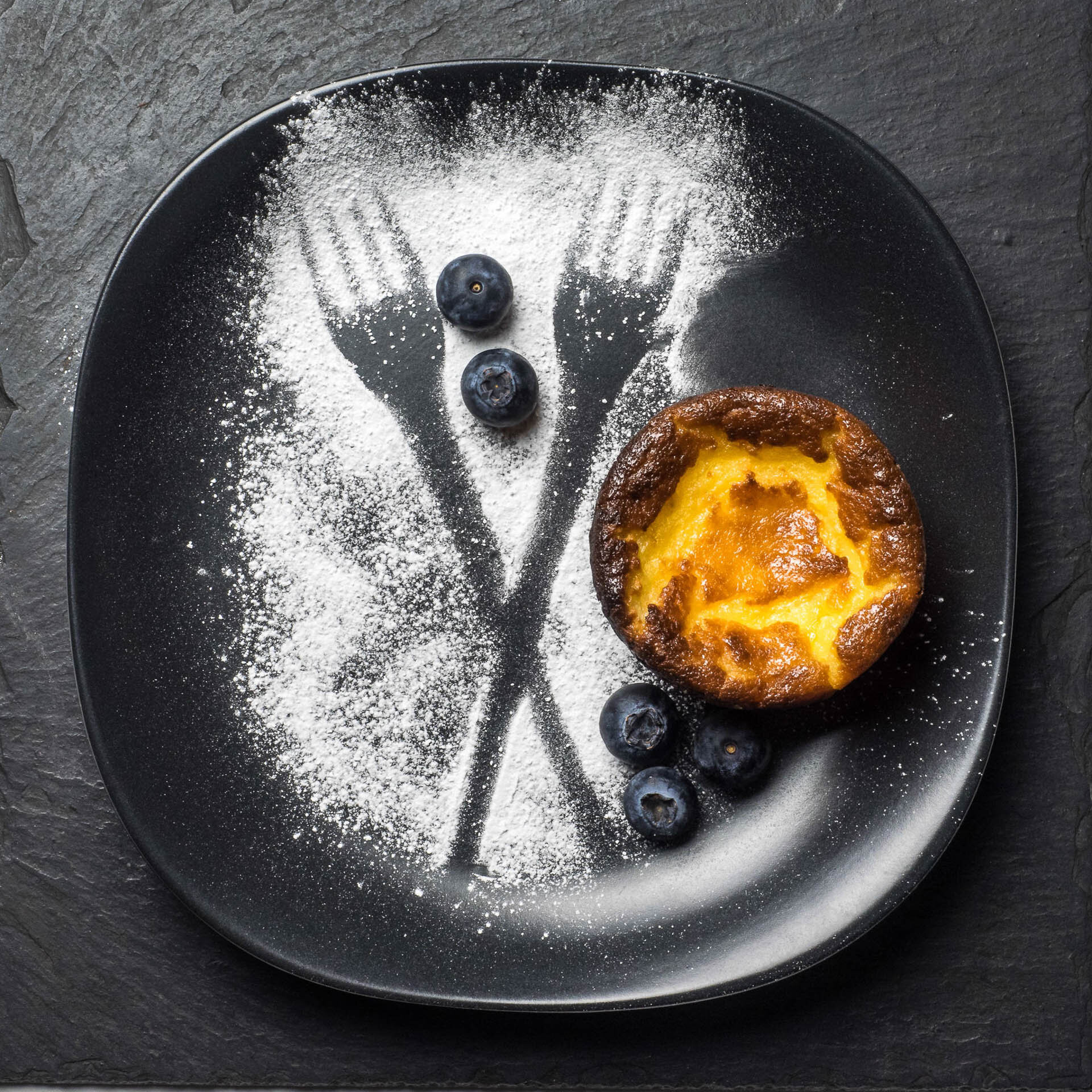 A black plate with powdered sugar in the shape of crossed forks, topped with blueberries, next to a baked muffin.