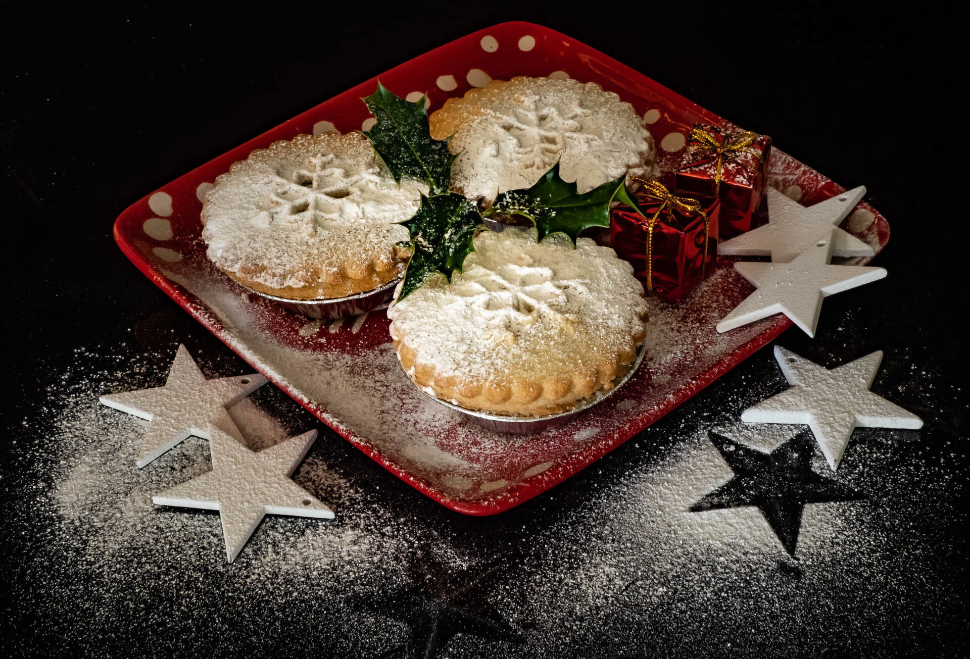 Three powdered sugar-dusted mince pies with holly leaves on a red plate, accompanied by small decorative gift boxes and star ornaments on a black background.