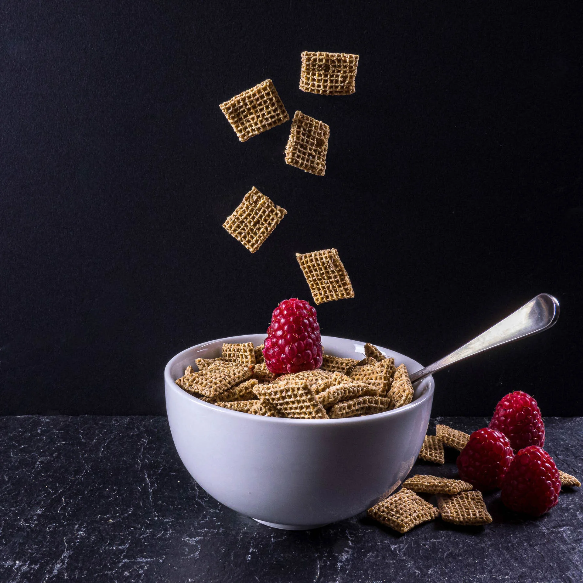 A bowl of cereal with a raspberry on top, surrounded by floating cereal pieces and additional raspberries on a dark surface.