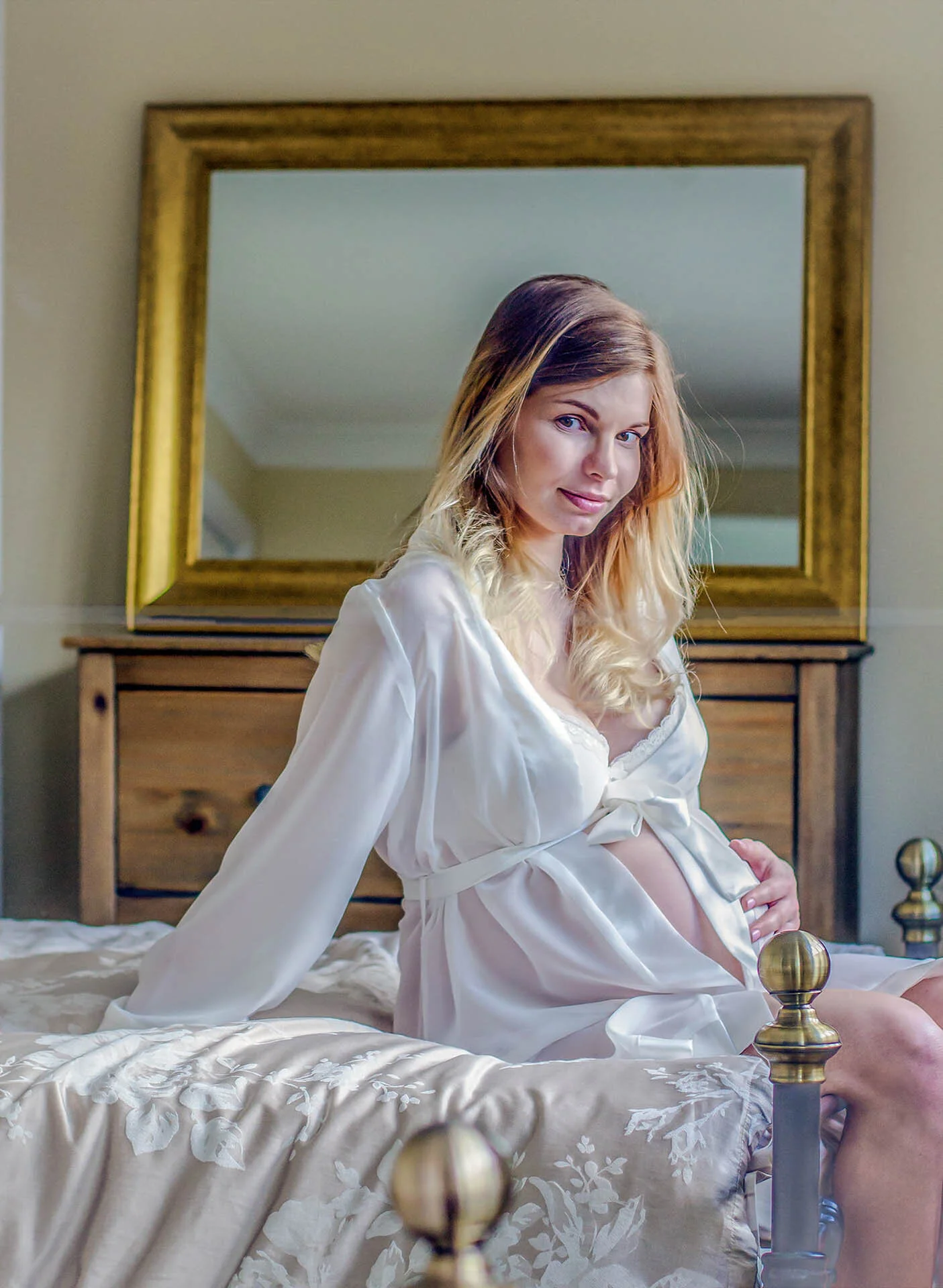 Pregnant woman in white robe sitting on a bed with floral bedding, with a wooden dresser and mirror in the background.