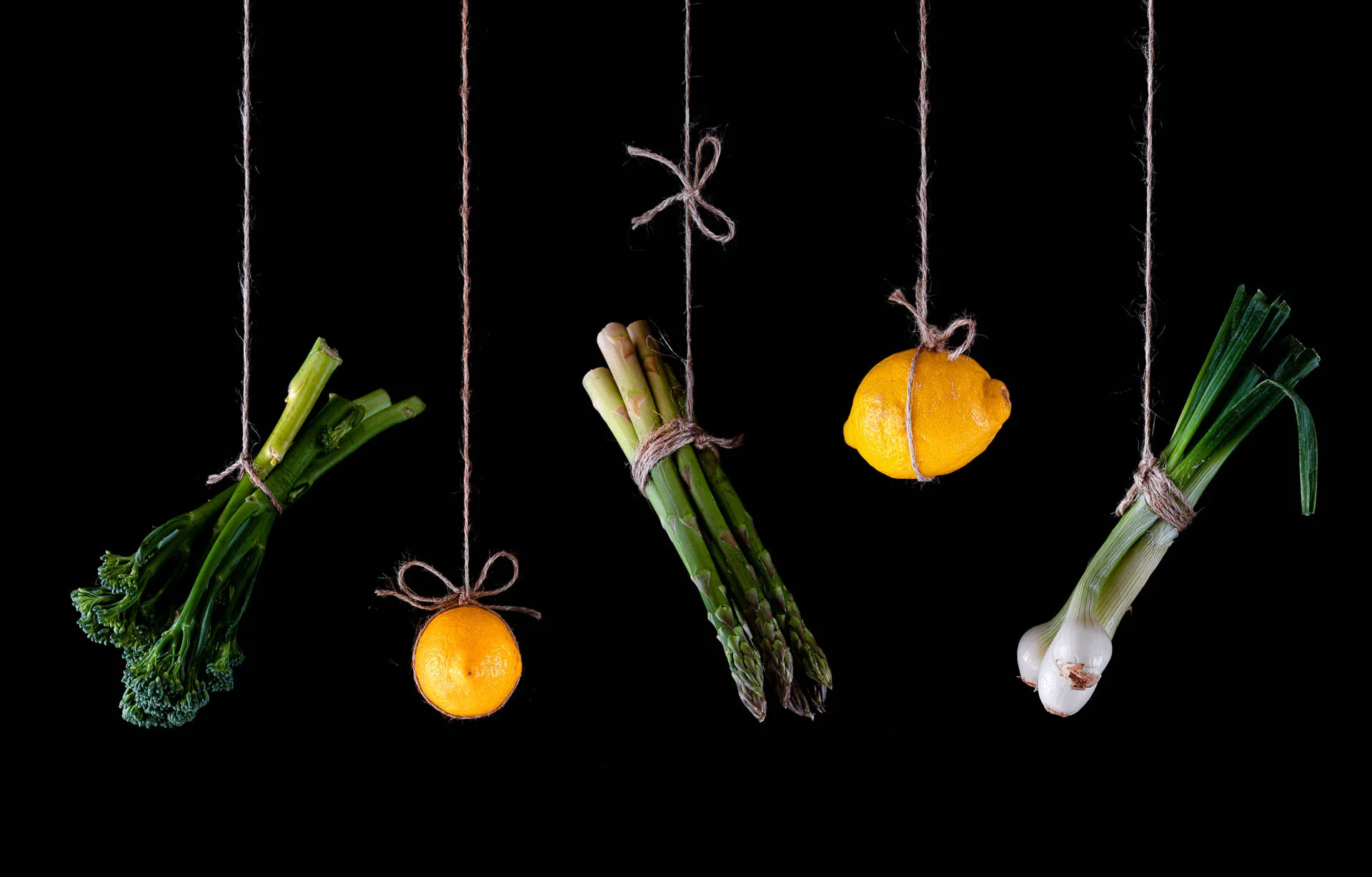 Vegetables and fruits hanging on strings against a black background, including broccolini, orange, asparagus, lemon, and green onions.