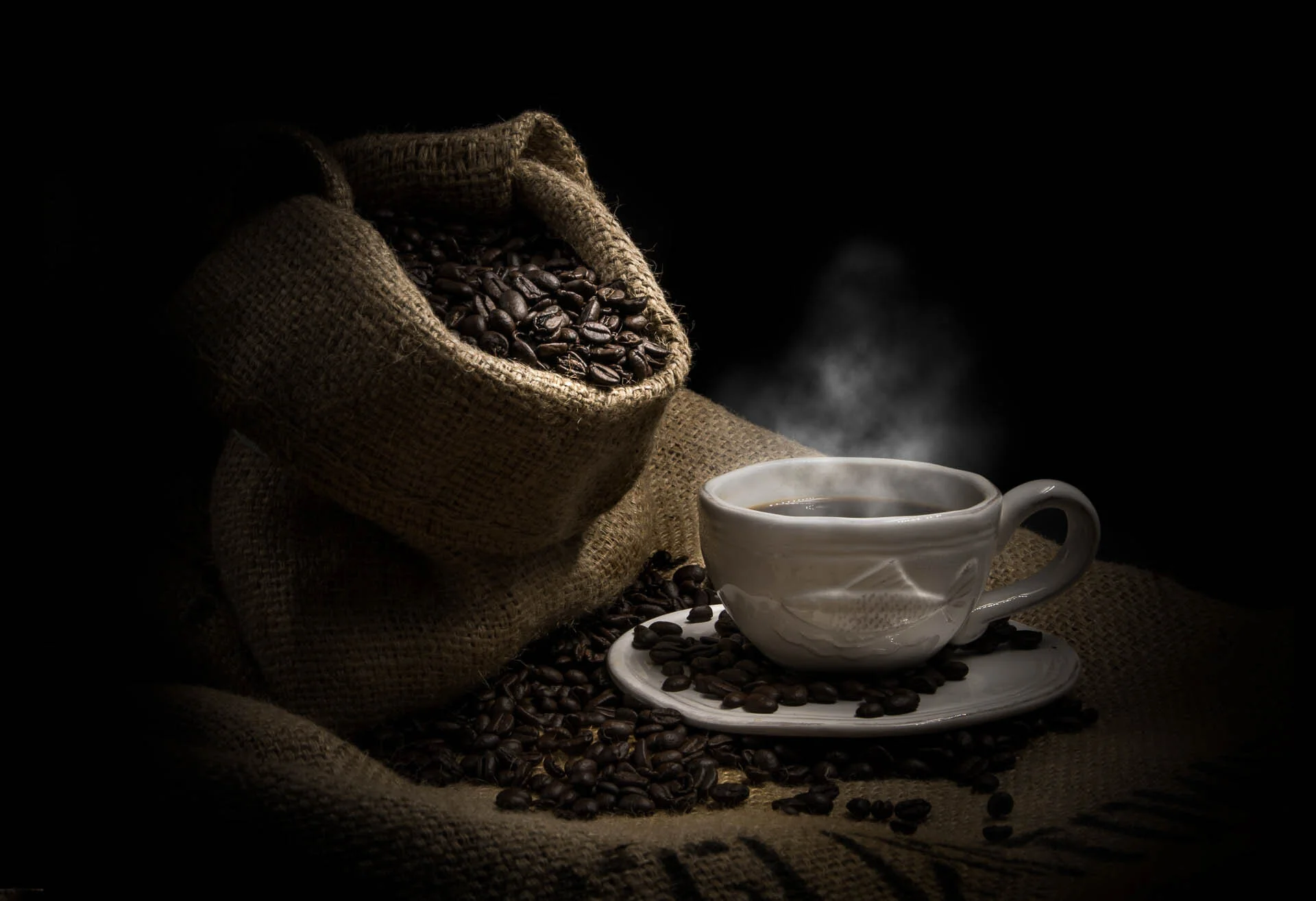 Steaming cup of coffee on saucer with burlap sack filled with coffee beans.