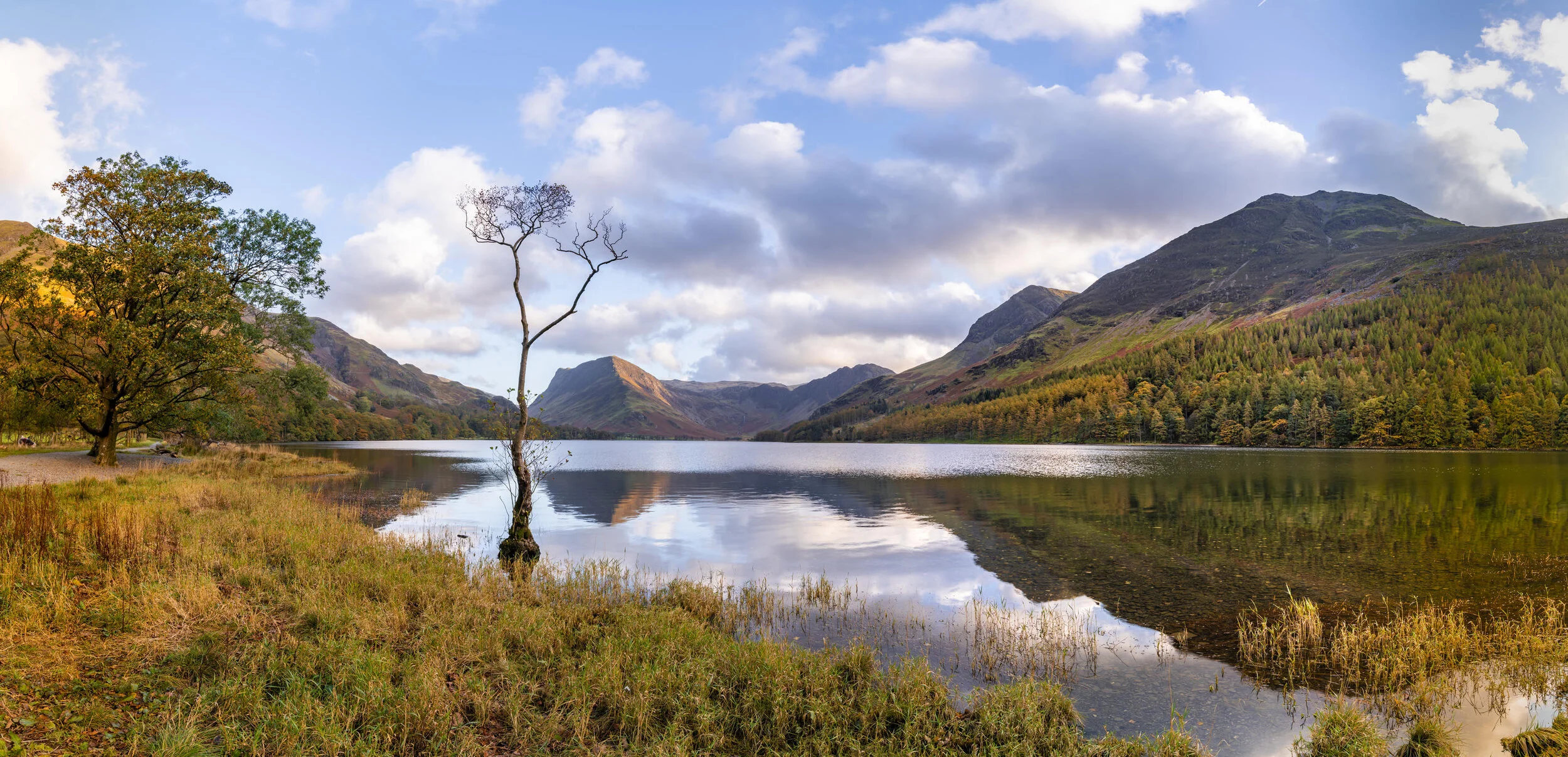 Scenic view of a lake surrounded by mountains and trees with reflections on the water, under a partly cloudy sky.