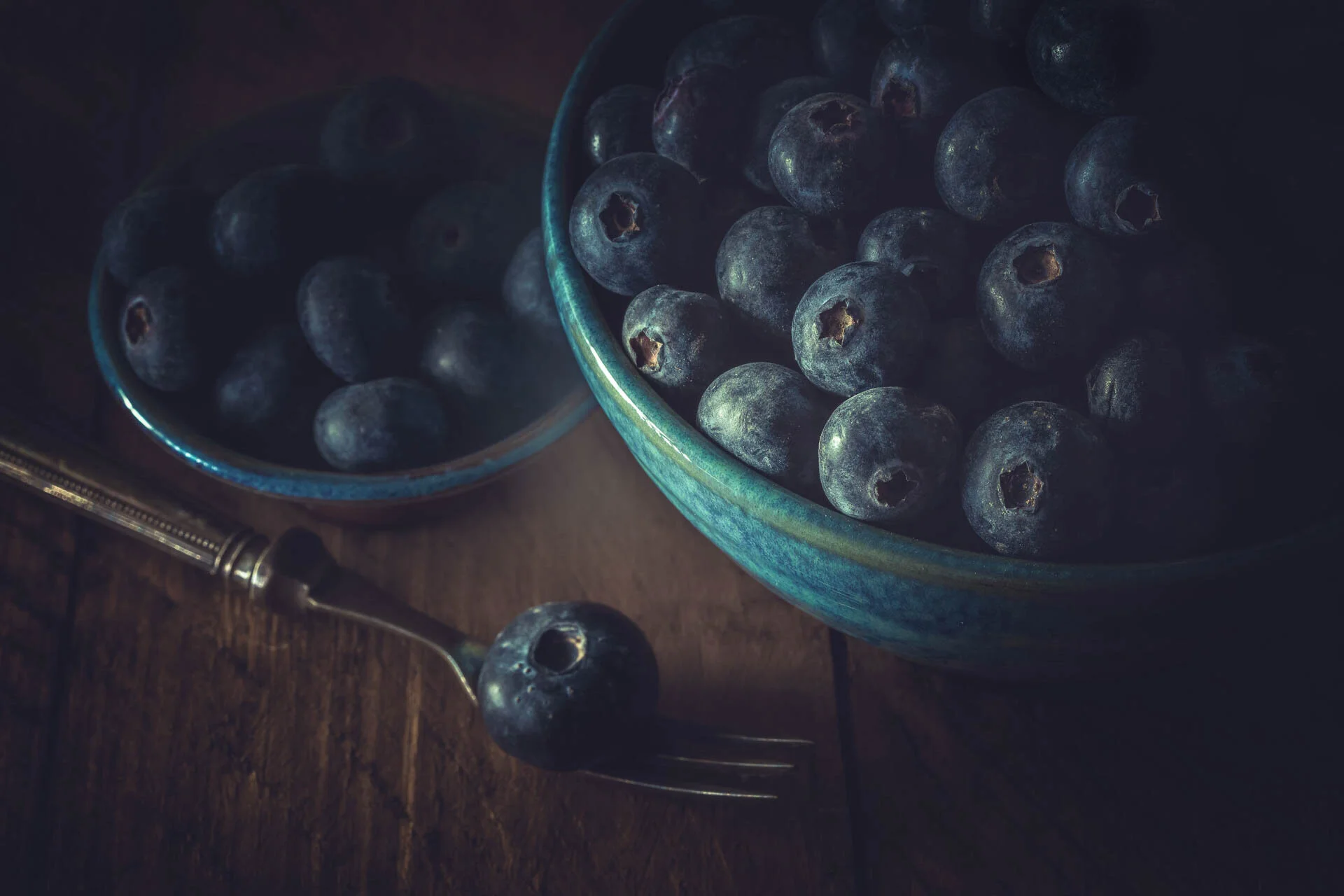 Close-up of blueberries in two ceramic bowls, with one blueberry on a fork, on a wooden table.